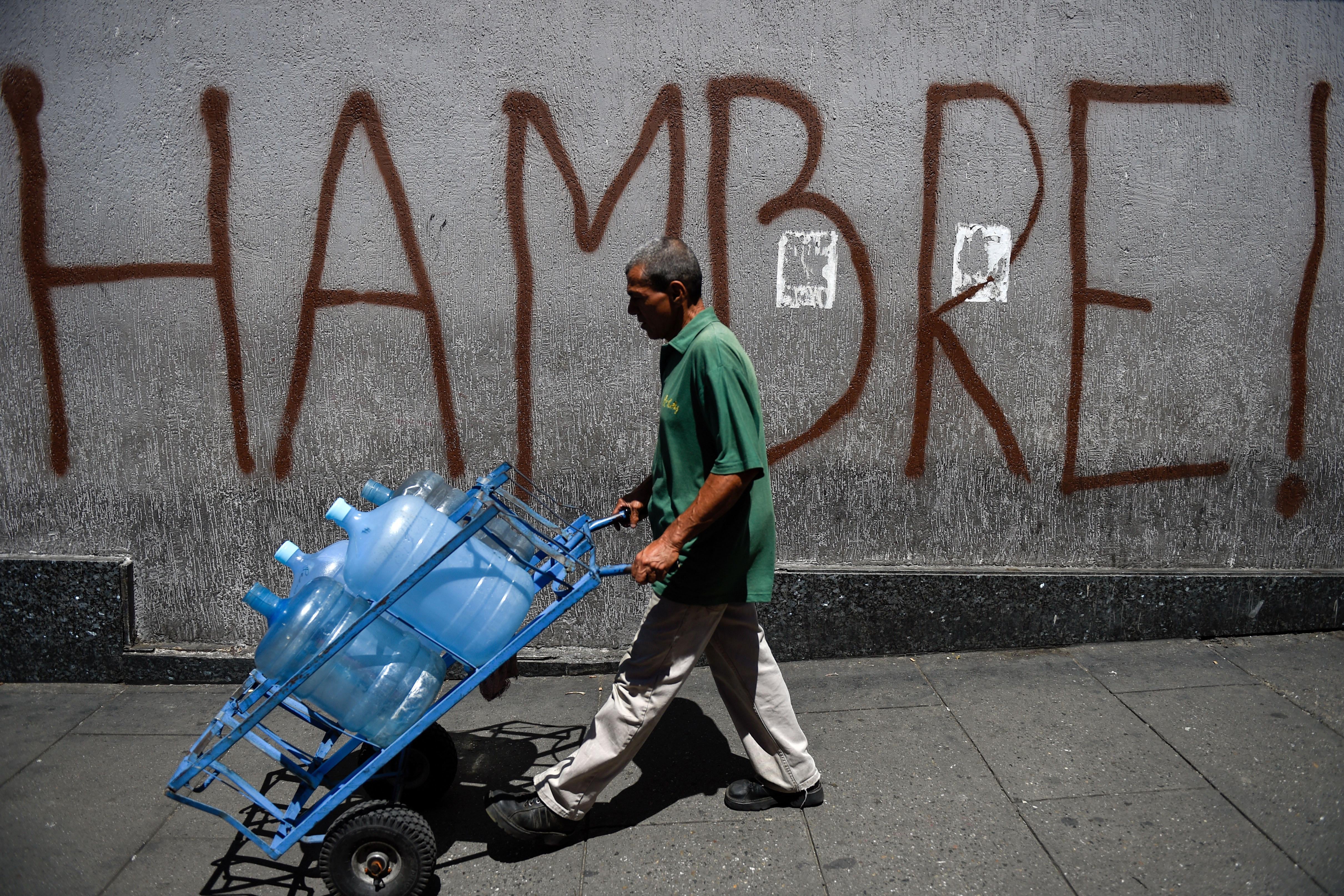 A Venezuelan man pushes a hand truck loaded with water jugs in front of street graffiti reading "HAMBRE!" or "hungry" in Spanish.