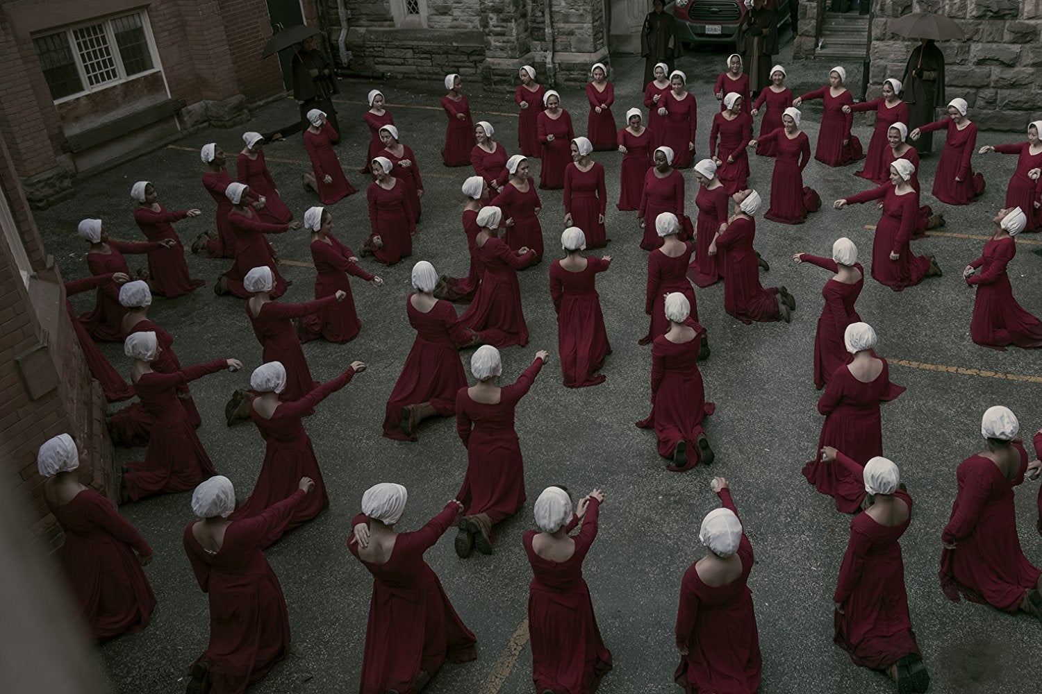 View from above: Women wearing red robes and white bonnets kneel in concentric circles, each with one arm outstretched.