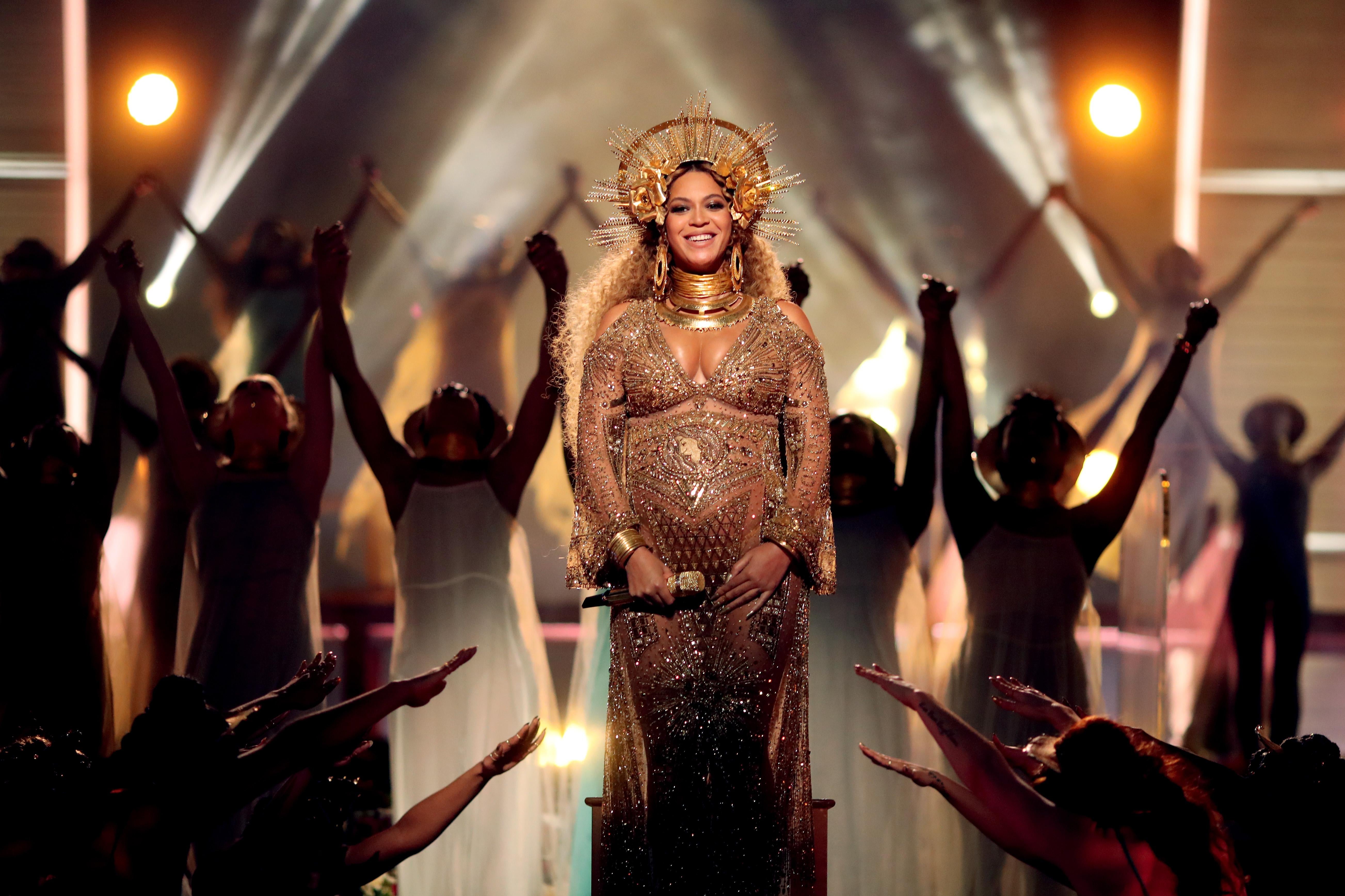 Beyonce standing on stage smiling in a long sleeve gold dress and a halo crown.