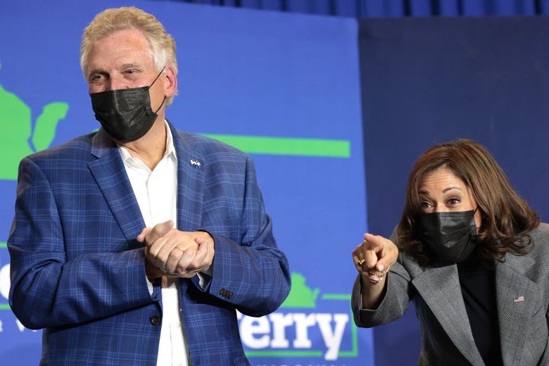 McAuliffe and Harris, both wearing masks and blazers, smile onstage. Harris is bending over and pointing animatedly toward someone in the audience.