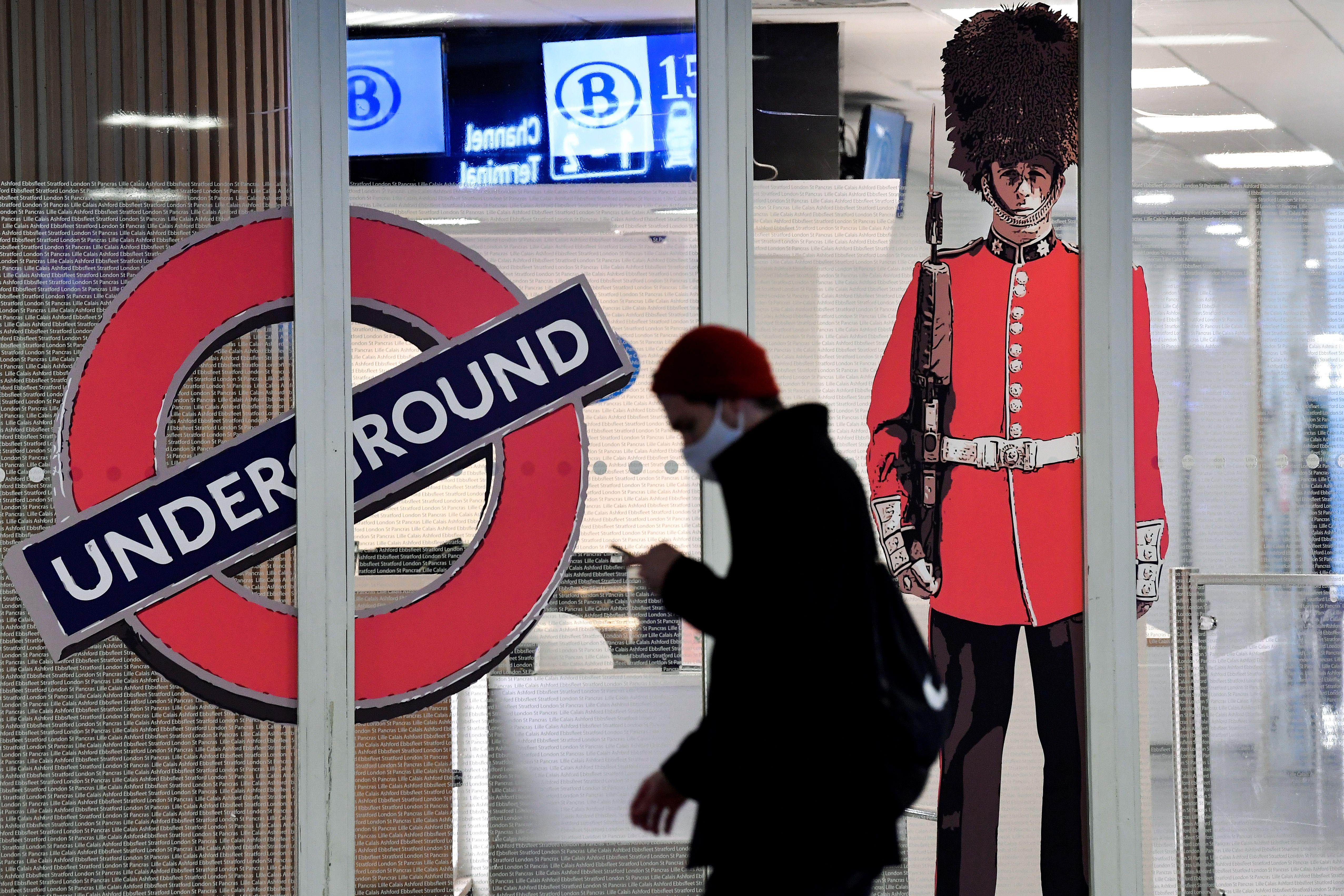A person wearing a mask and looking at their phone walks past a London Underground sign and a Queen's Guard standee at the closed entrance of the Eurostar terminal at Brussels South railway station.