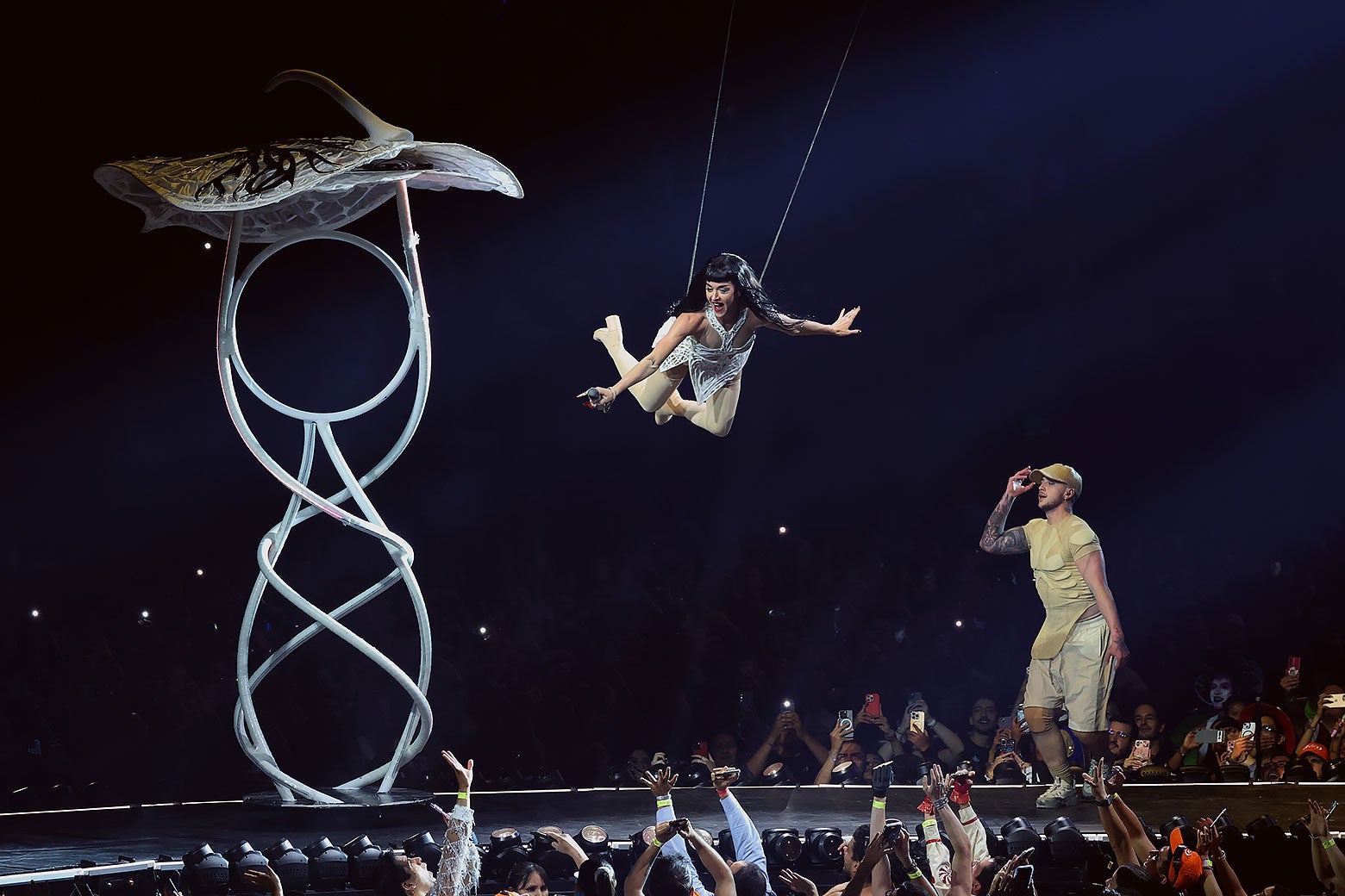 Perry wears a white costume and hangs suspended above a stage.