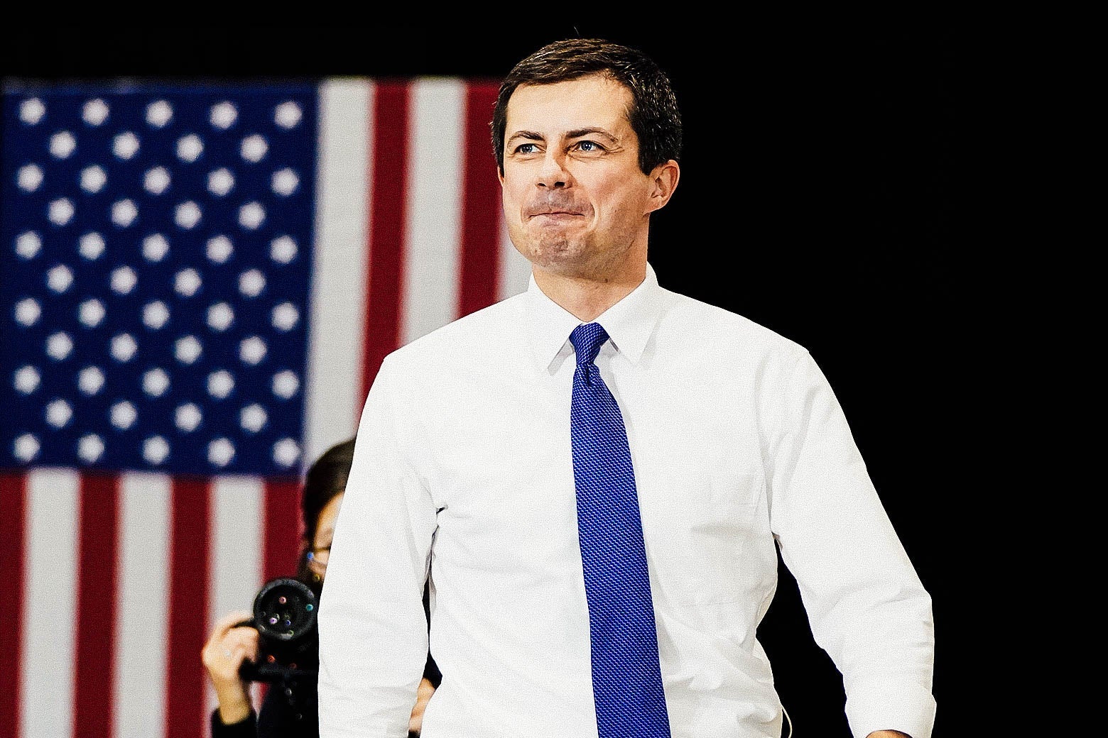 Buttigieg smiling as he walks, with an American flag in the background.