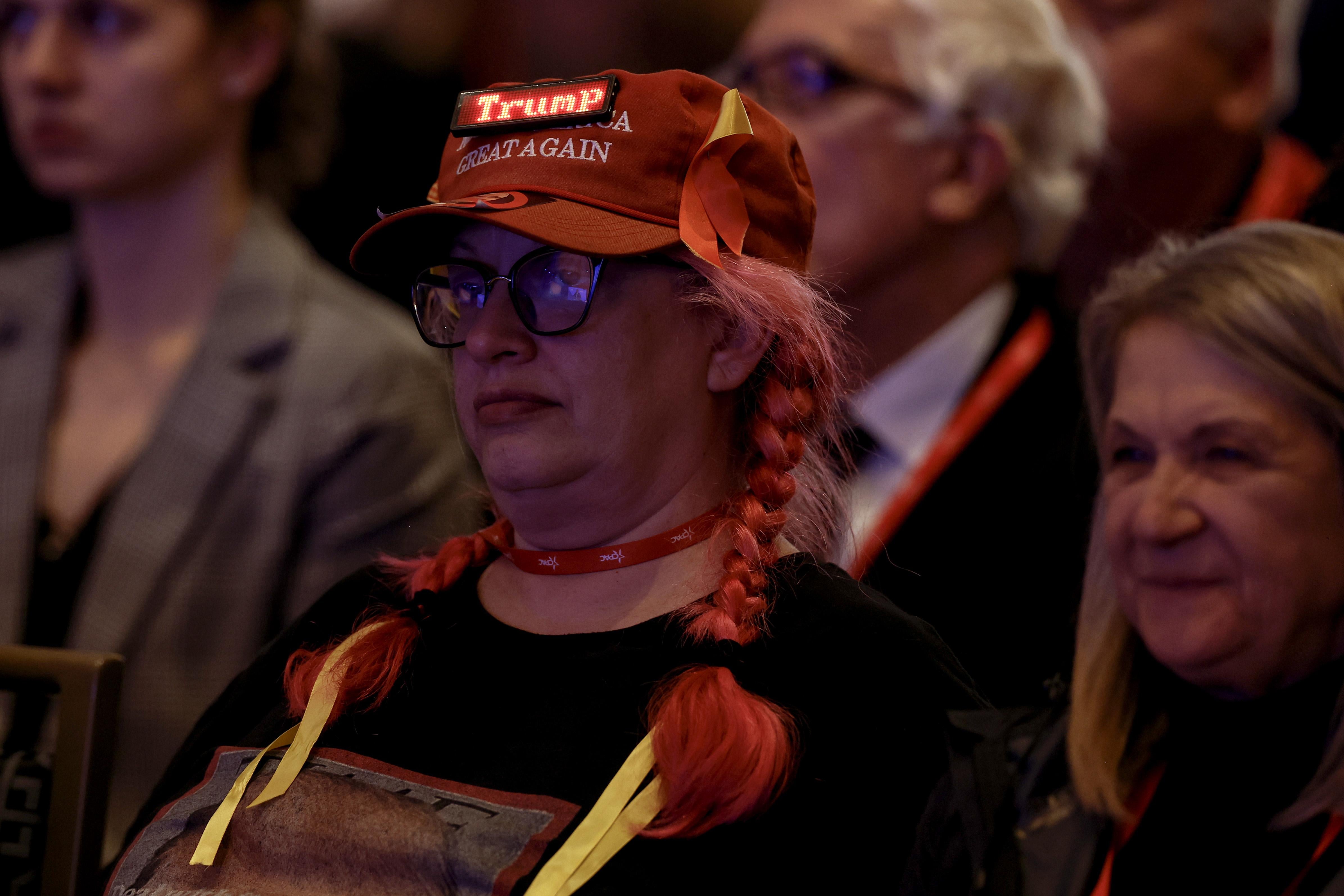 An adult woman wears a MAGA hat with some kind of Thor earflaps and Thor pigtails. A Trump patch is adhered to the MAGA hat. The woman frowns.