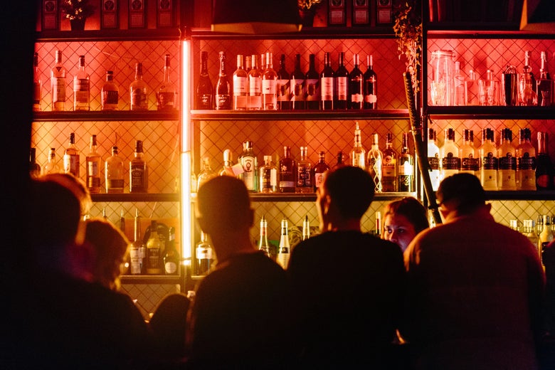 A bar with bottles of alcohol. We can see backs of the heads of several young men who are sitting at the bar drinking. 