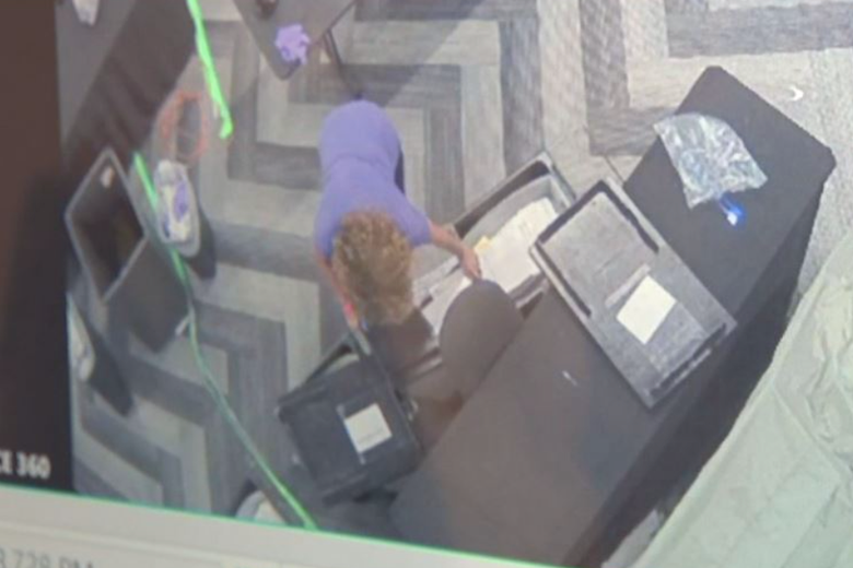 A woman leans over what is very clearly a crate filled with papers in a still image from security camera video taken from an overhead angle.