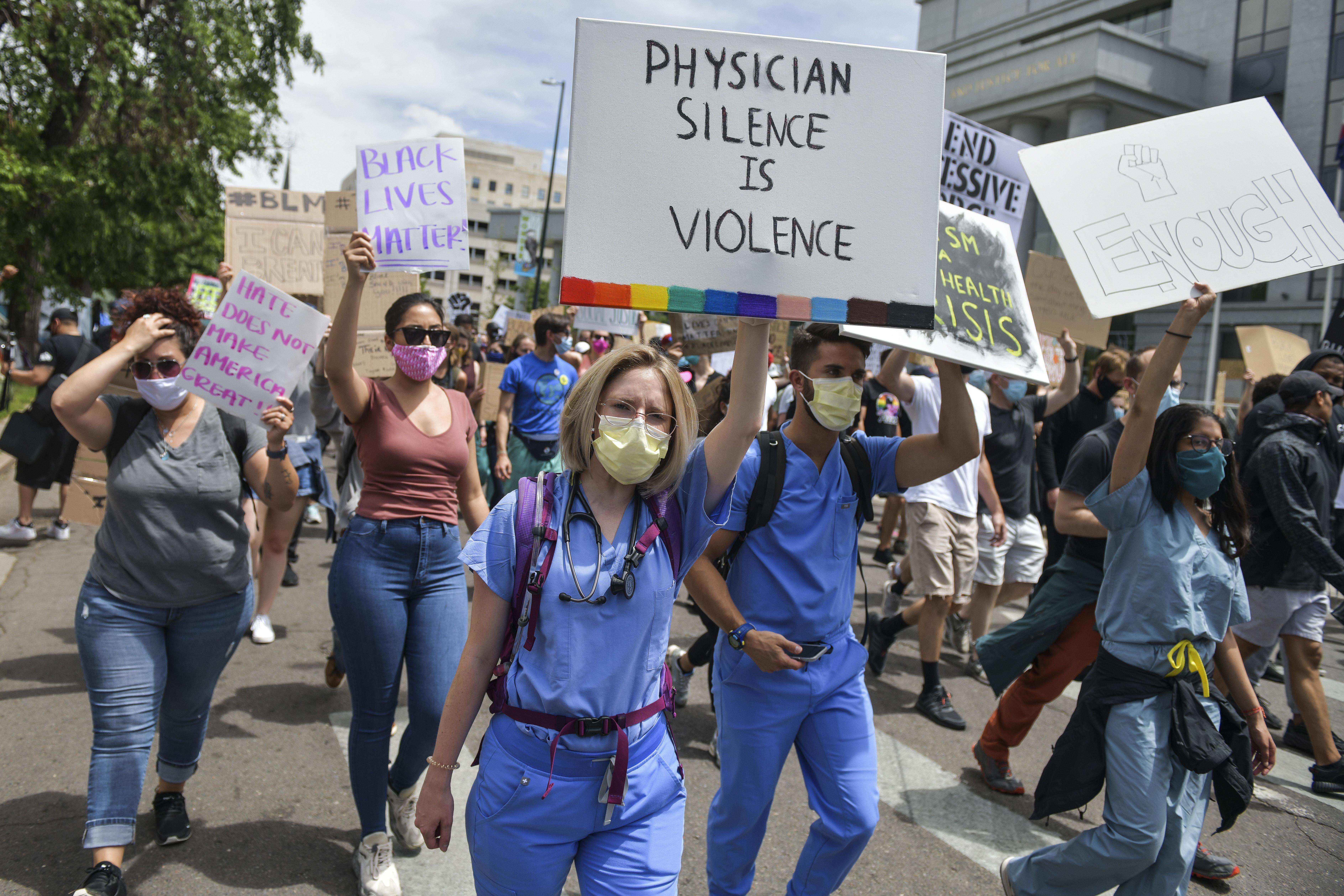 Physicians in scrubs march with other Black Lives Matter protesters. One physician holds a sign that says "Physician silence is violence."