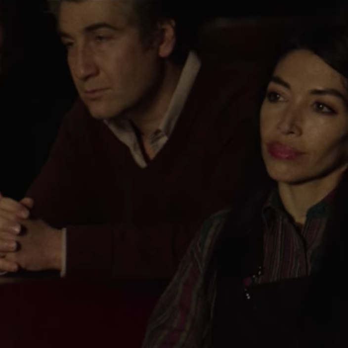 A photo of a clean-shaven man with brown hair sitting in the dark of a theater, in a collared shirt with a sweater over it