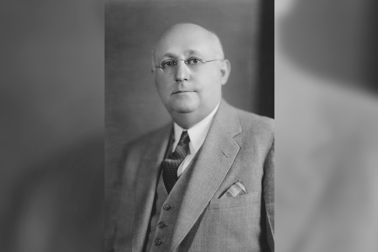 A white 1930s-era man: bald, wire-framed glasses, a light-gray suit and vest and black tie.