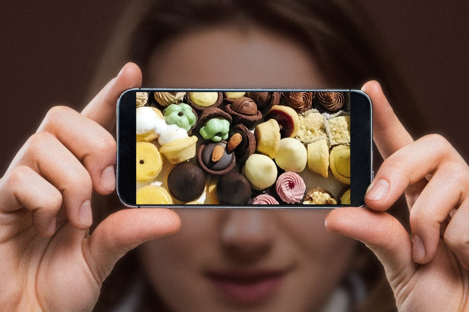 A young woman holds an iPhone horizontally in front of her face, with the screen facing the camera. On the phone screen is a photo of small pastries and a mix of bite-sized tarts in different flavors, with brown, green, pink, and white frosting. The phone is blocking the woman's eyes, but she has a slight smile and appears happy. 