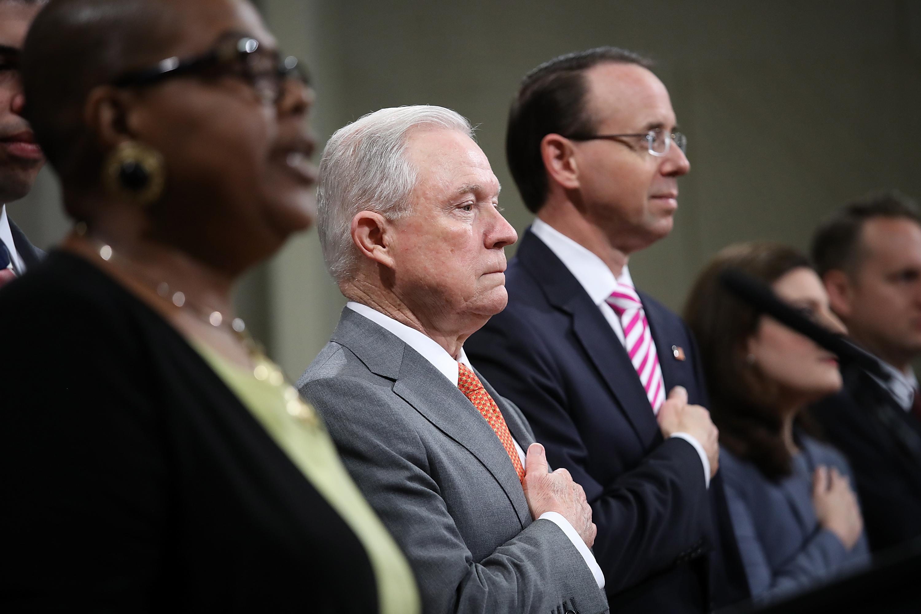 Jeff Sessions, standing to the left of Deputy Attorney General Rod Rosenstein, places his hand over his heart.