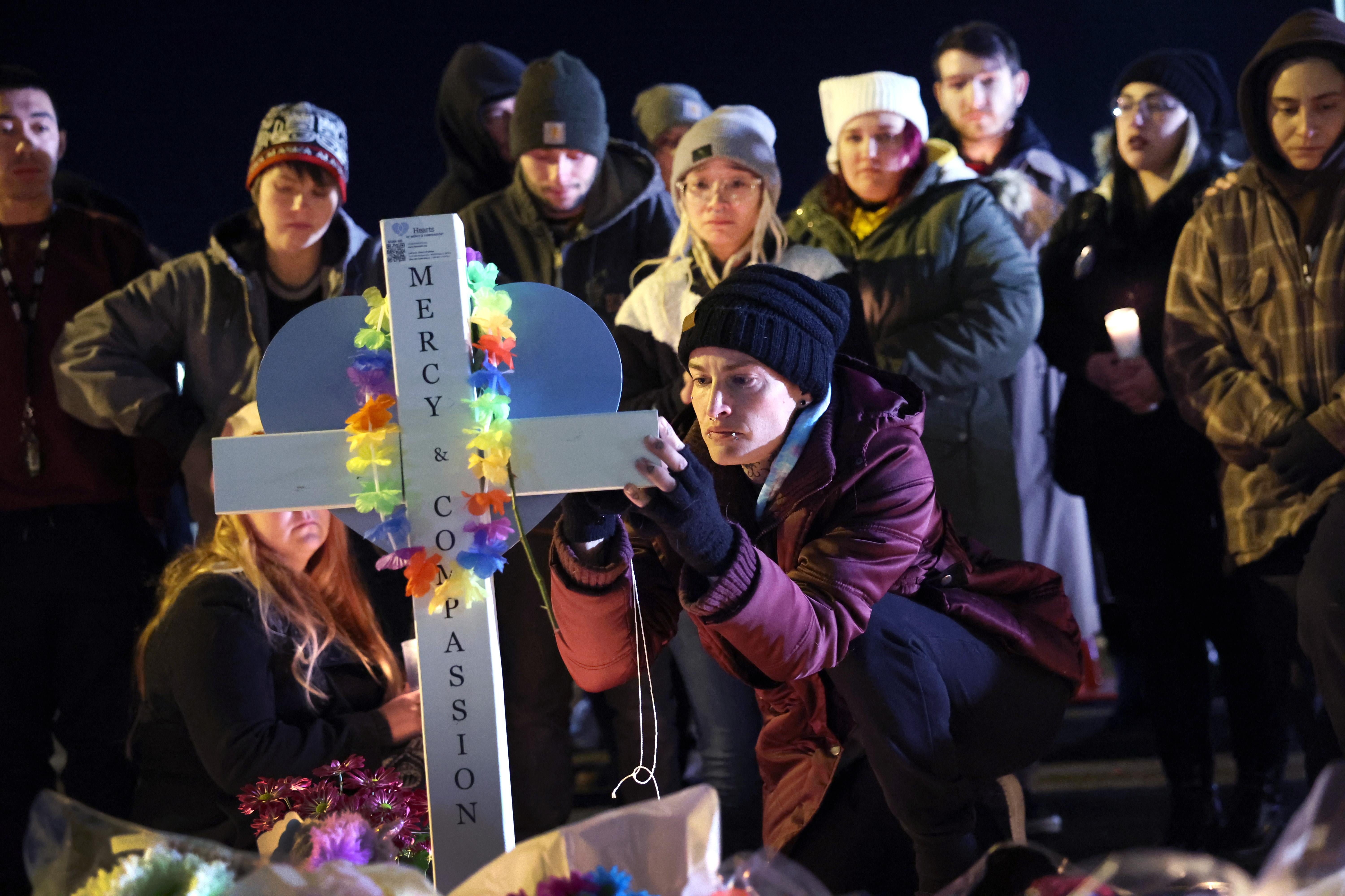 Mourners gather around a white cross with a rainbow-colored lei on it in the dark.