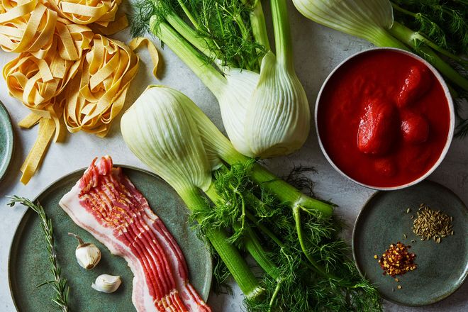 Fennel bulbs, uncooked pasta, tomato sauce, raw bacon, and various spices arranged on a kitchen countertop.