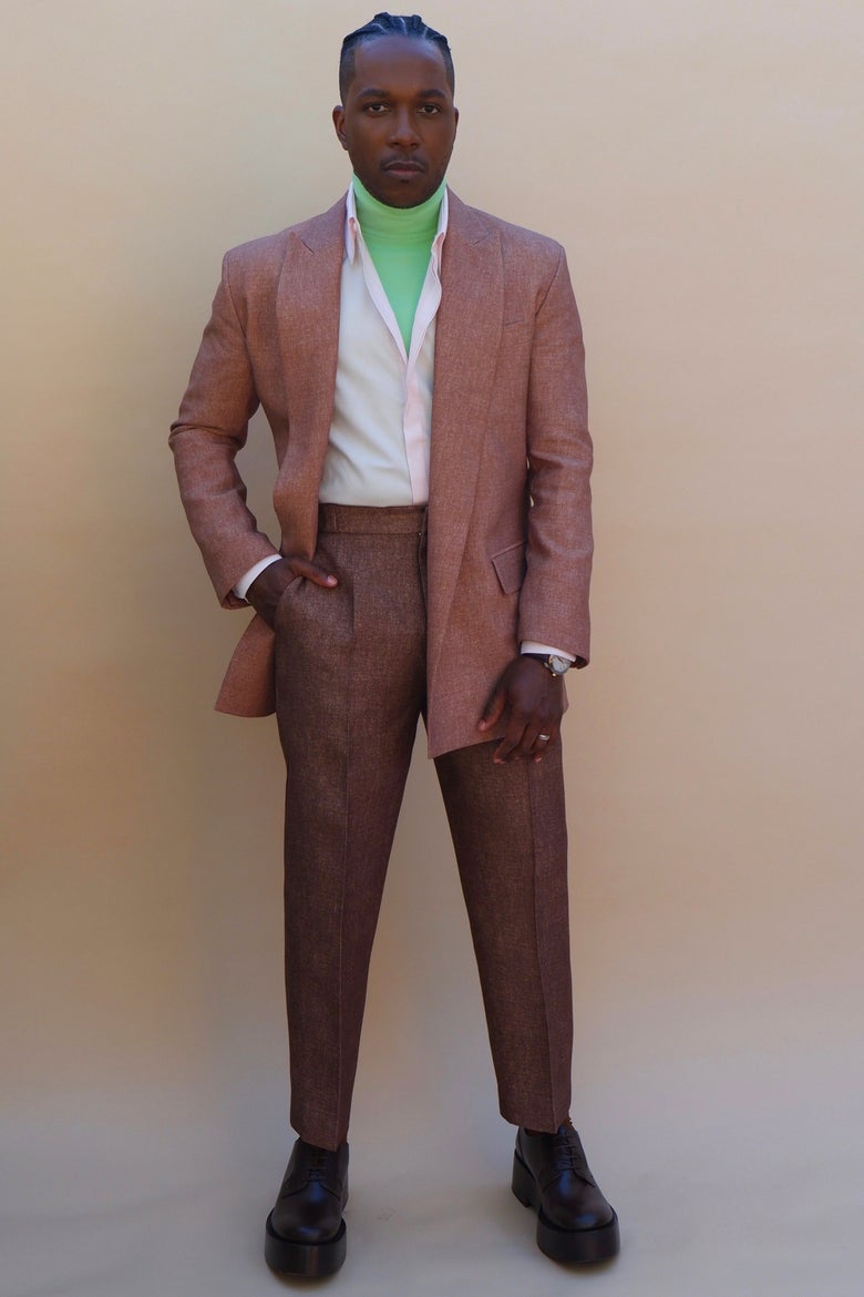 Leslie Odom stands in front of a peach background with one hand in his pants pocket.