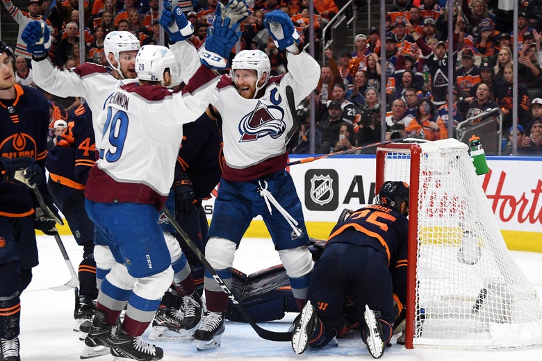 MacKinnon and teammates raise their arms and cheer in celebration around the goal, as Oilers frown and crouch, dejected, around them.