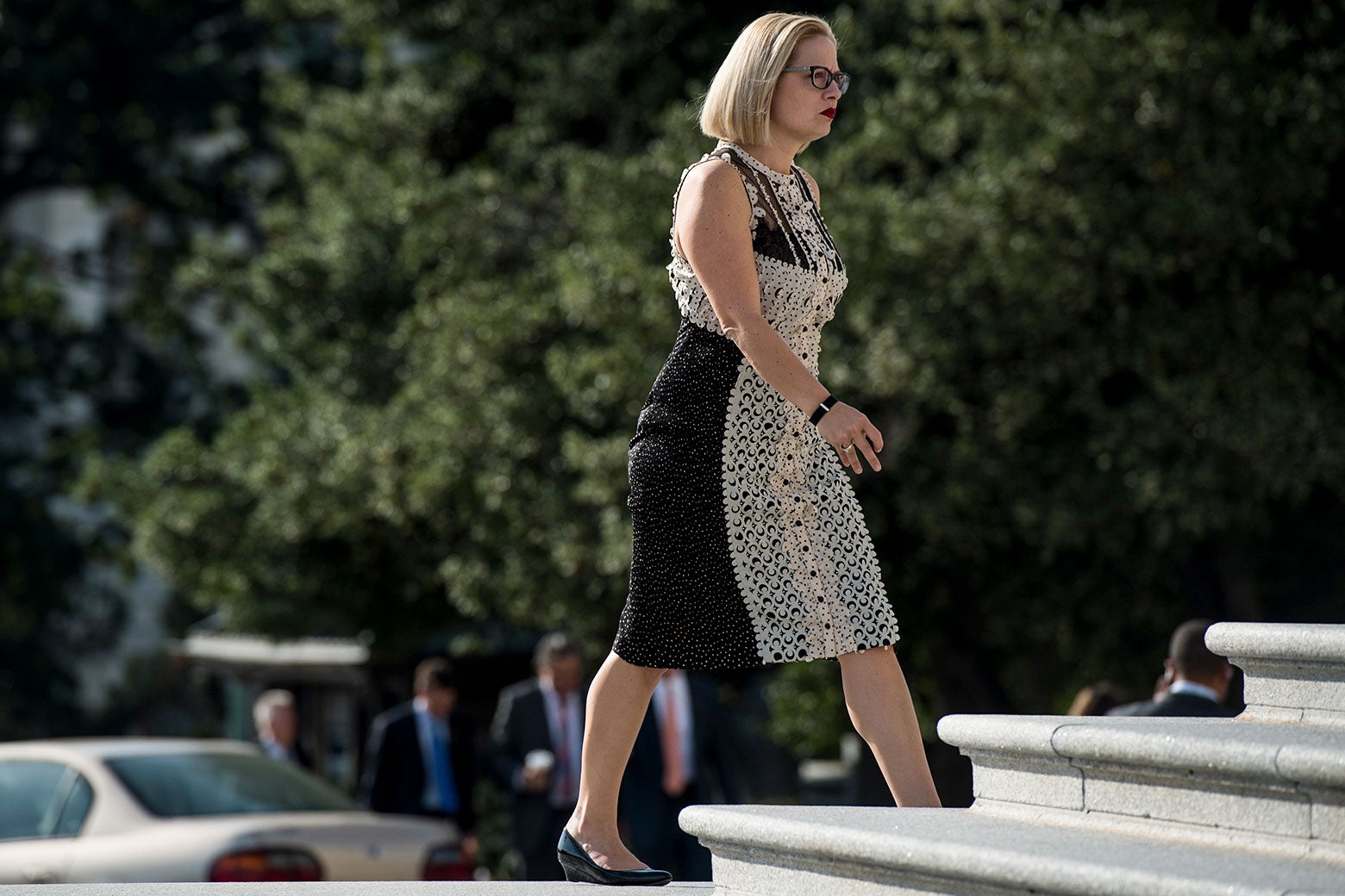 Kyrsten Sinema walks up the House steps in a textured, lacy sleeveless black and cream dress.