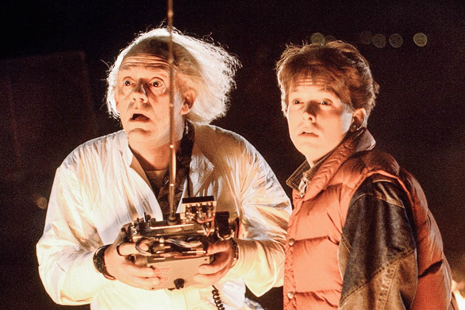 Doc Brown, with his big white hair blown back, holds a giant remote control next to Michael J. Box, in his puffy vest, in Back to the Future