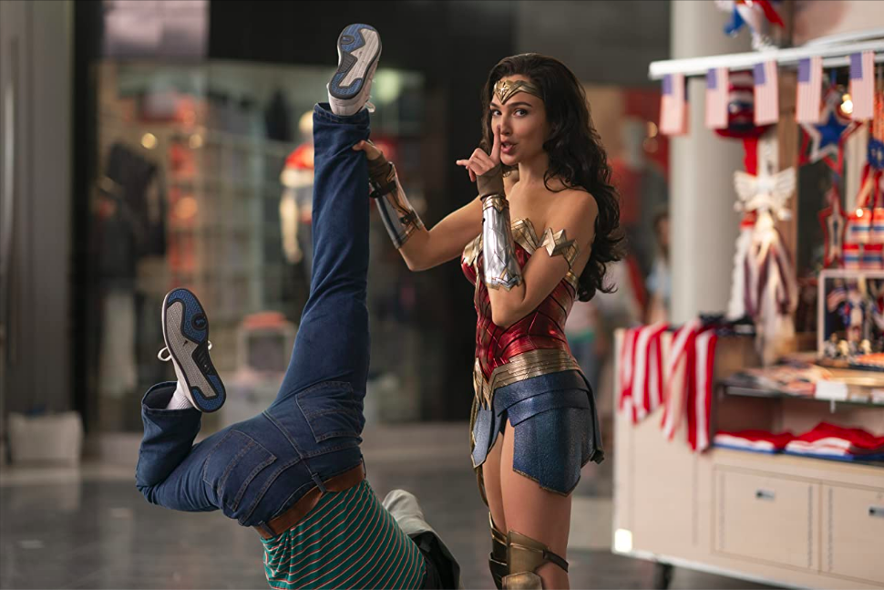 Wonder Woman, standing next to a kiosk in a mall, holds some poor sap upside-down while turning to the camera and holding one finger to her lips