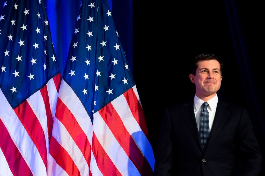 Buttigeg, wearing a suit, walks on to a stage in front of two American flags.