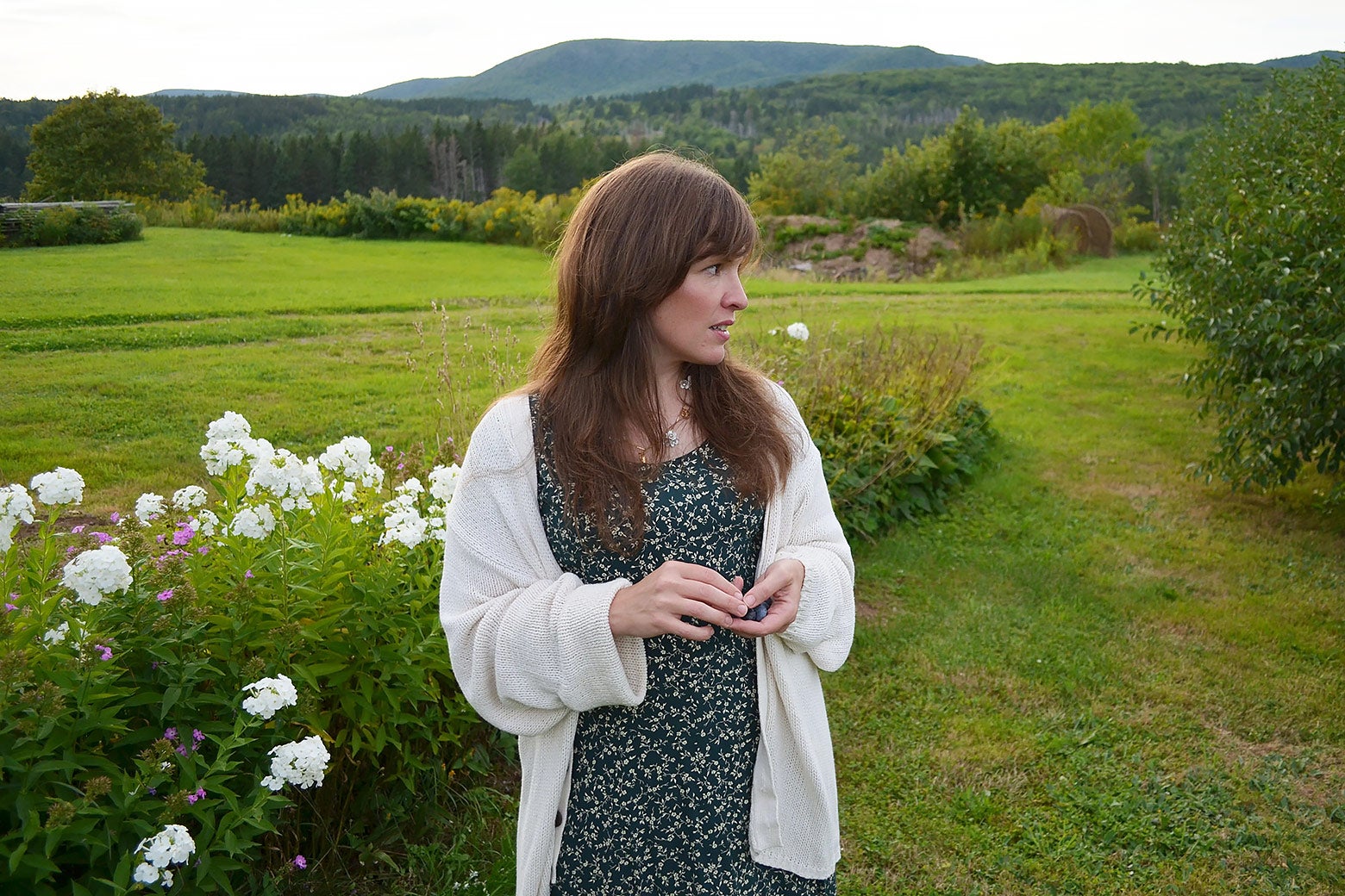 A woman wearing a dress stands in a field, looking to her right.