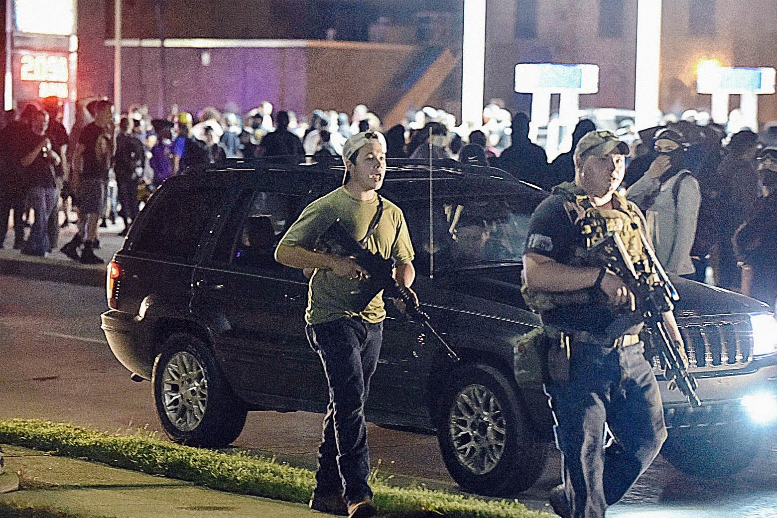Rittenhouse in a T-shirt holding an AR-style rifle walks with Balch in a bulletproof vest also holding an AR-style rifle at night. Behind them are a black SUV and a large crowd of people.