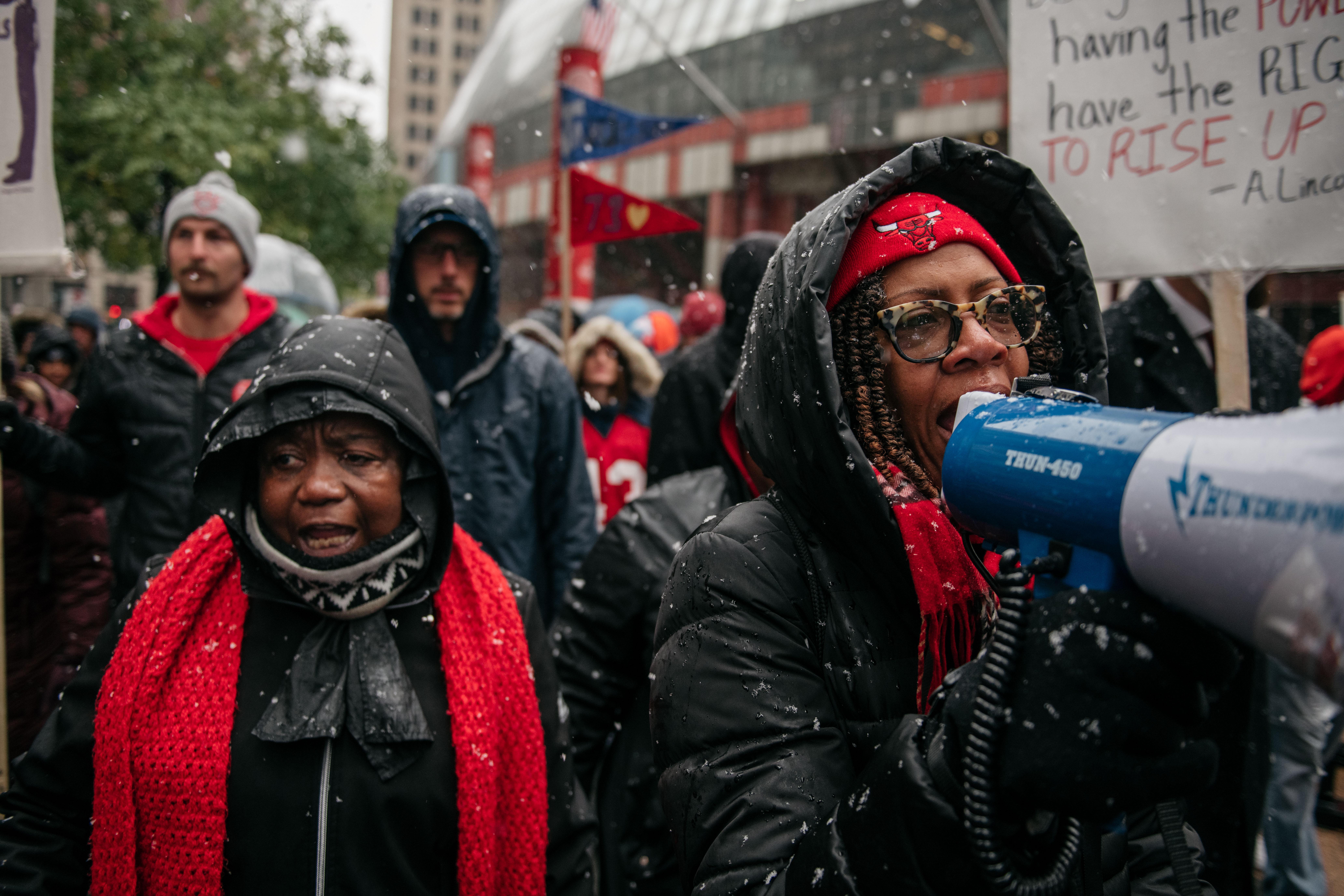 A woman in a crowd of demonstrators speaks into a megaphone. Some hold sings, and snow falls around the marchers.