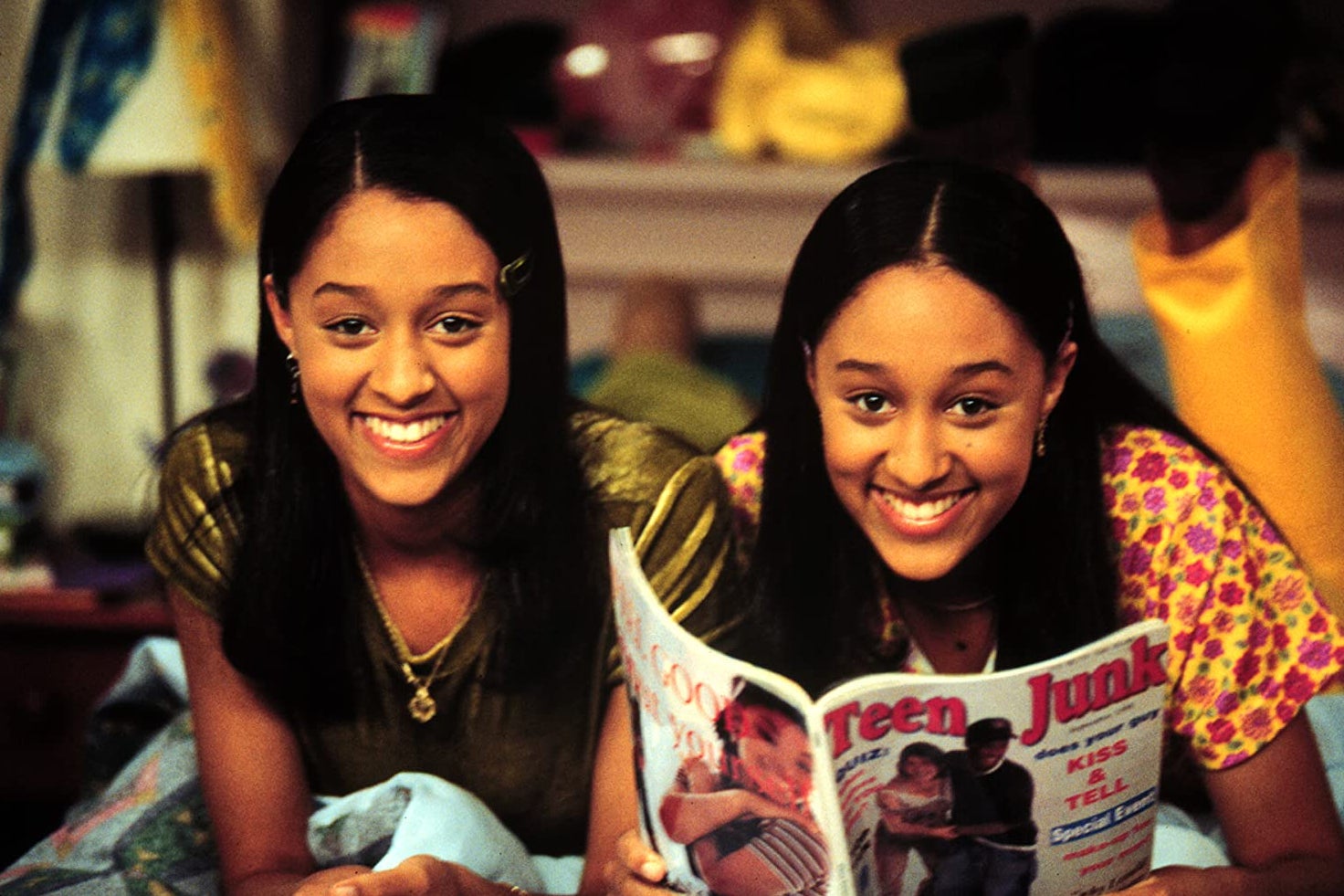 Tia and Tamera Mowry smiling and leaning forward on their elbows. Tamera holds a magazine with "Teen Junk" on the cover.