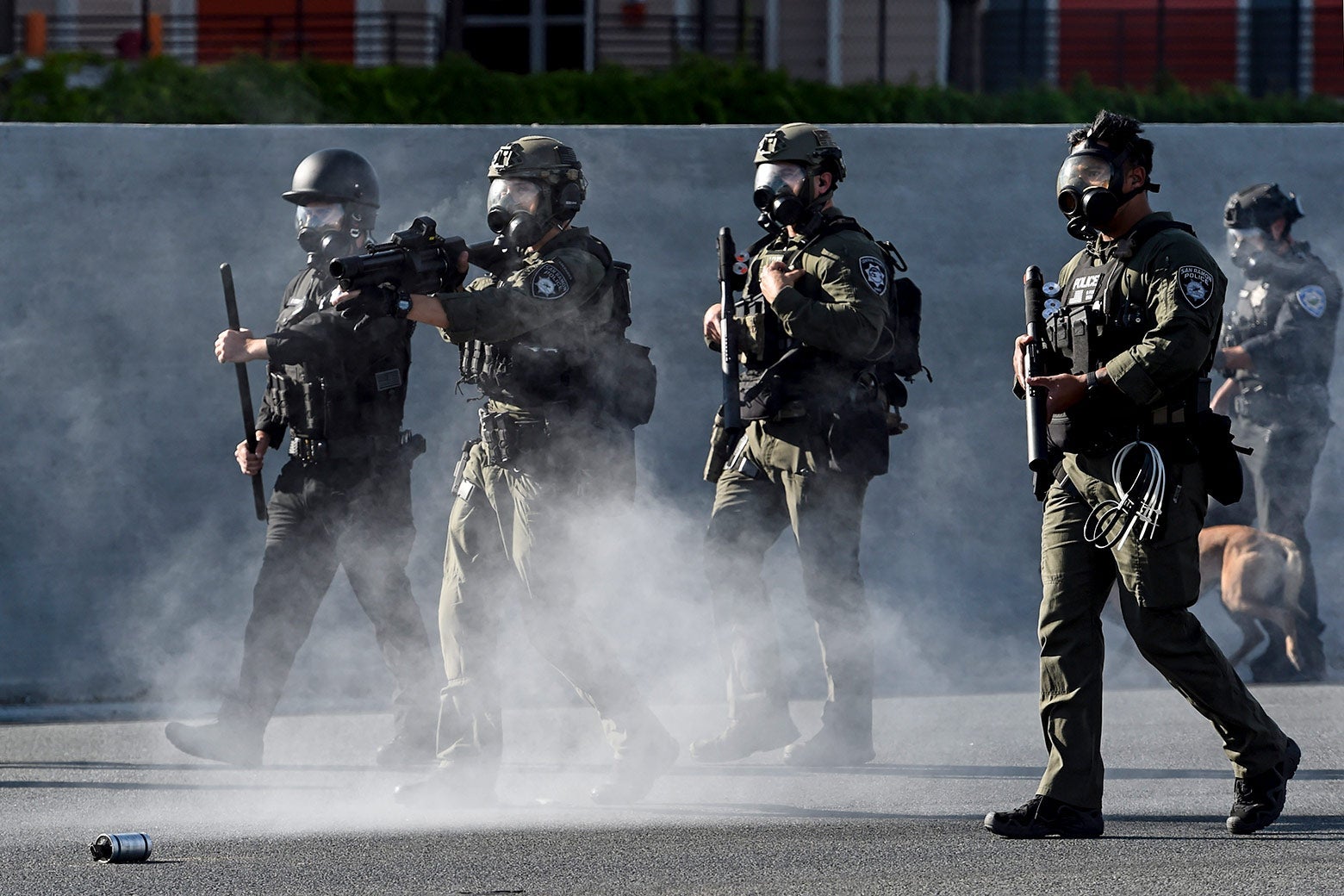 Police in tactical gear carry guns and throw tear-gas canisters.