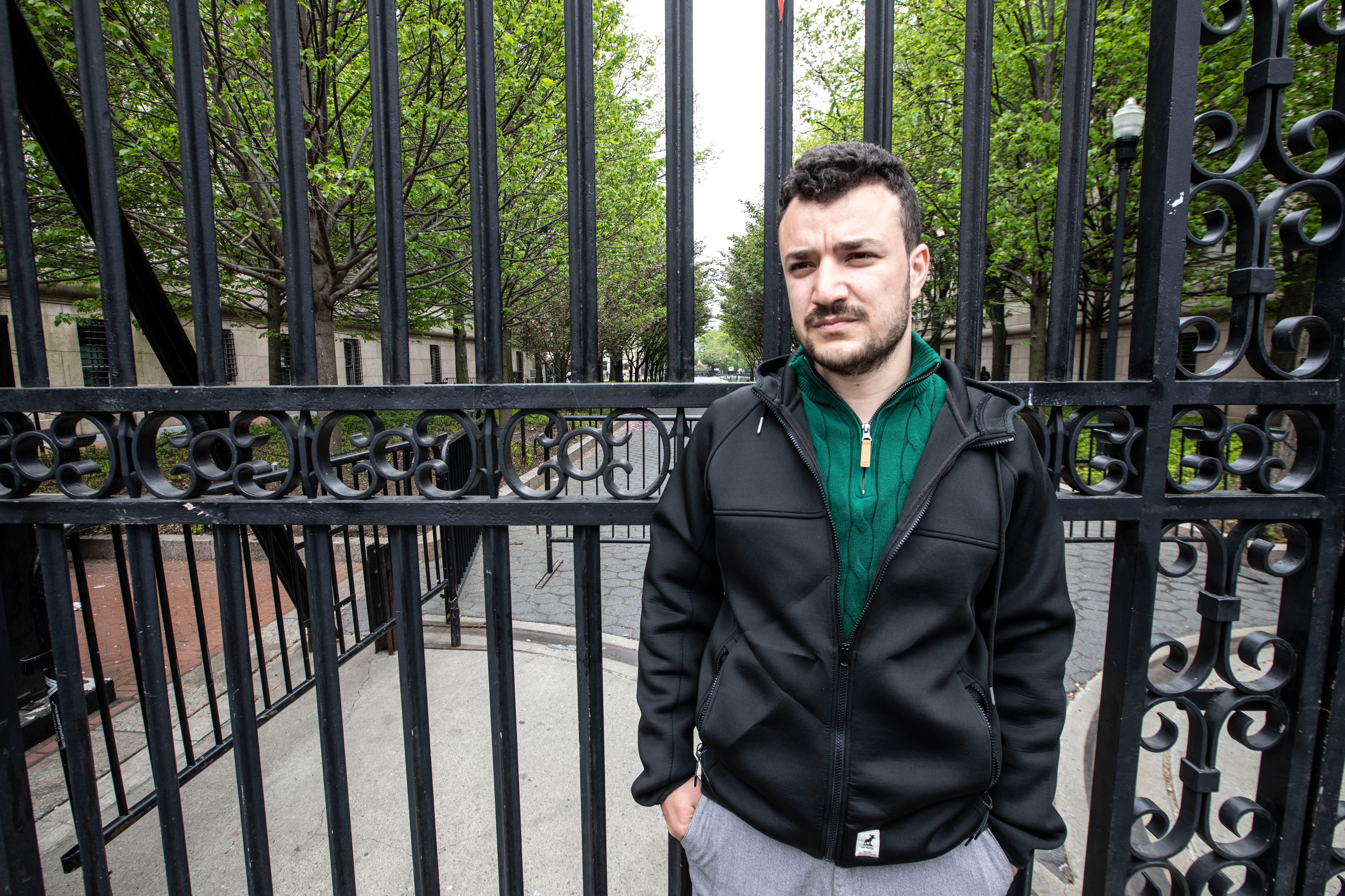 A young man with dark hair wears a green zip-up sweater and leather jacket and stands, his hands in his pockets, looking slightly serious.