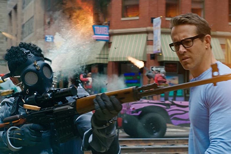 Ryan Reynolds, wearing a blue shirt, looks with confusion at a man in a gas mask firing a rifle.