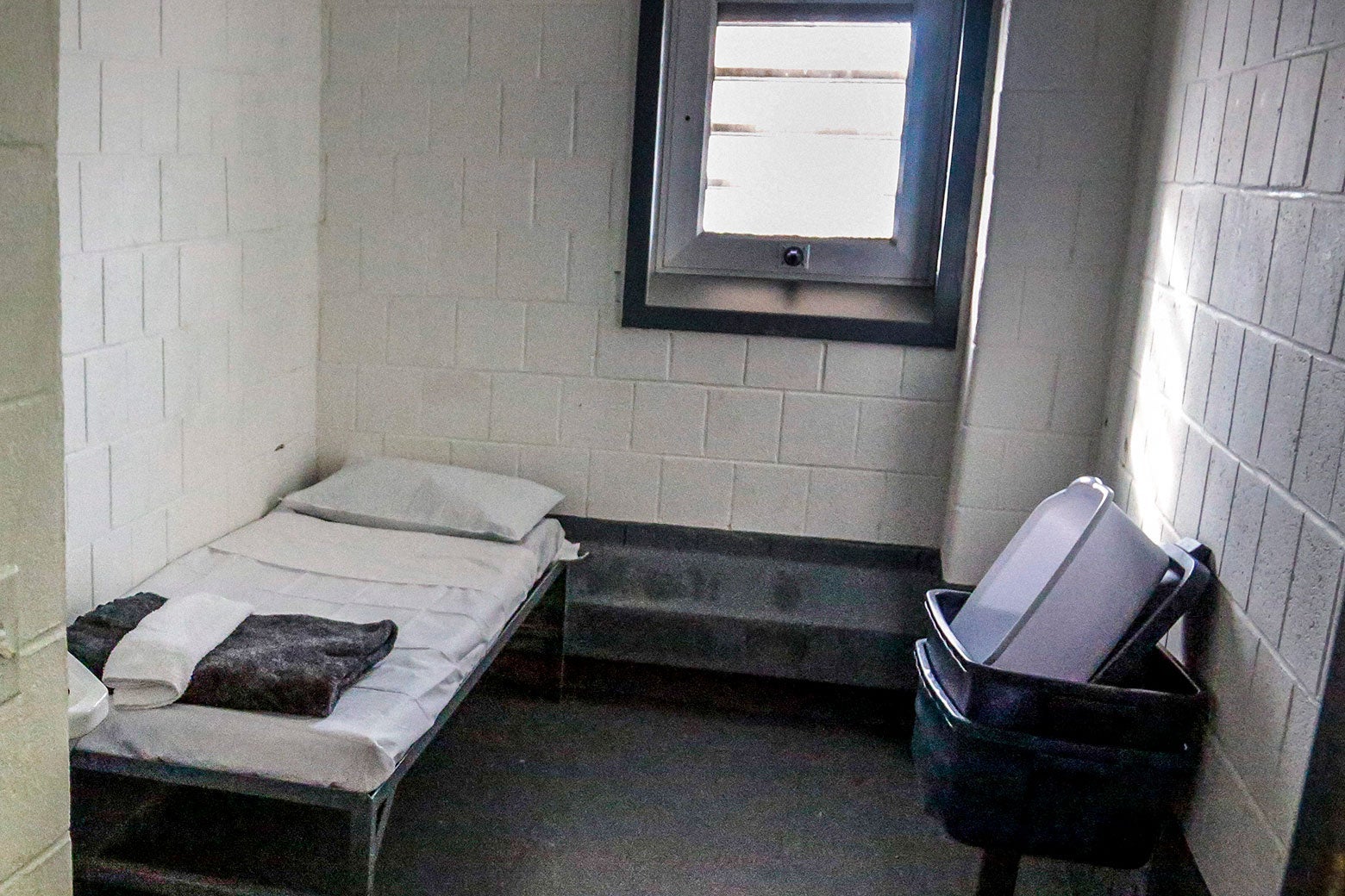 A tiny jail cell with a cot on one side and some plastic tubs on the other.