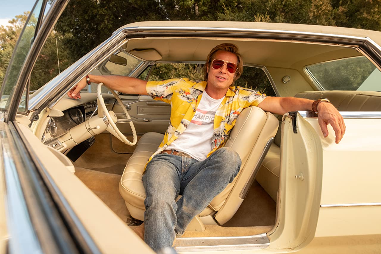 Brad Pitt, in costume as Cliff Booth in a still from Once Upon a Time in Hollywood, lounging in a cream colored Cadillac.