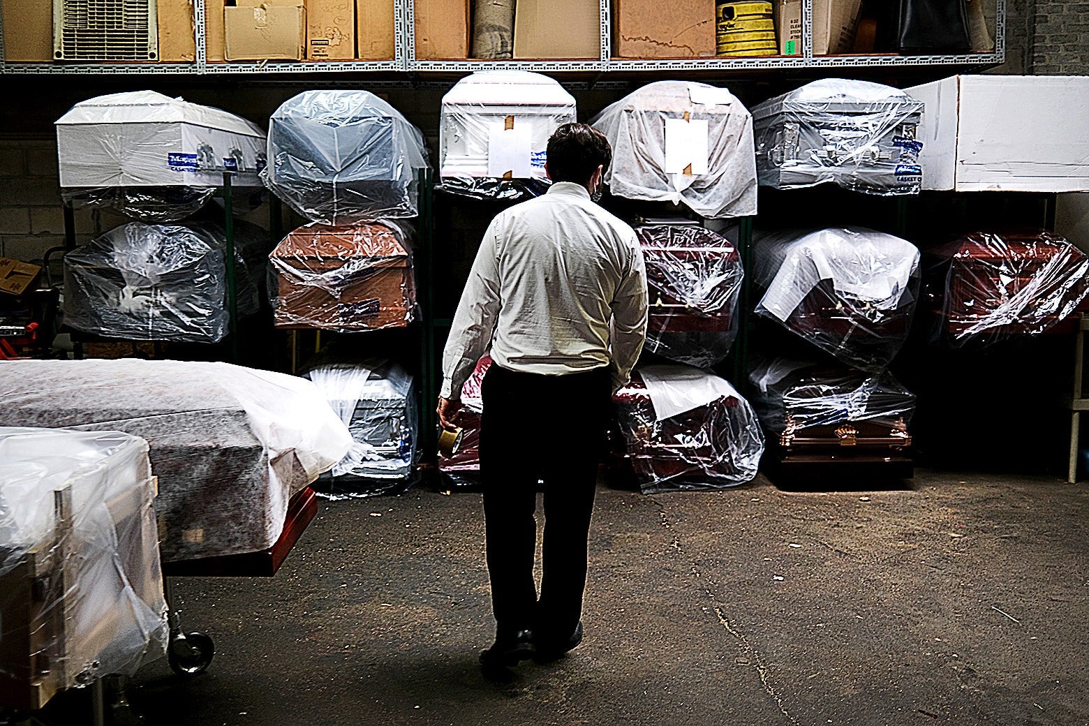 A man in a white shirt and dark pants stands with his back to the camera in front of a rack of coffins in various colors, individually wrapped in clear plastic sheeting, stacked three rows high. 