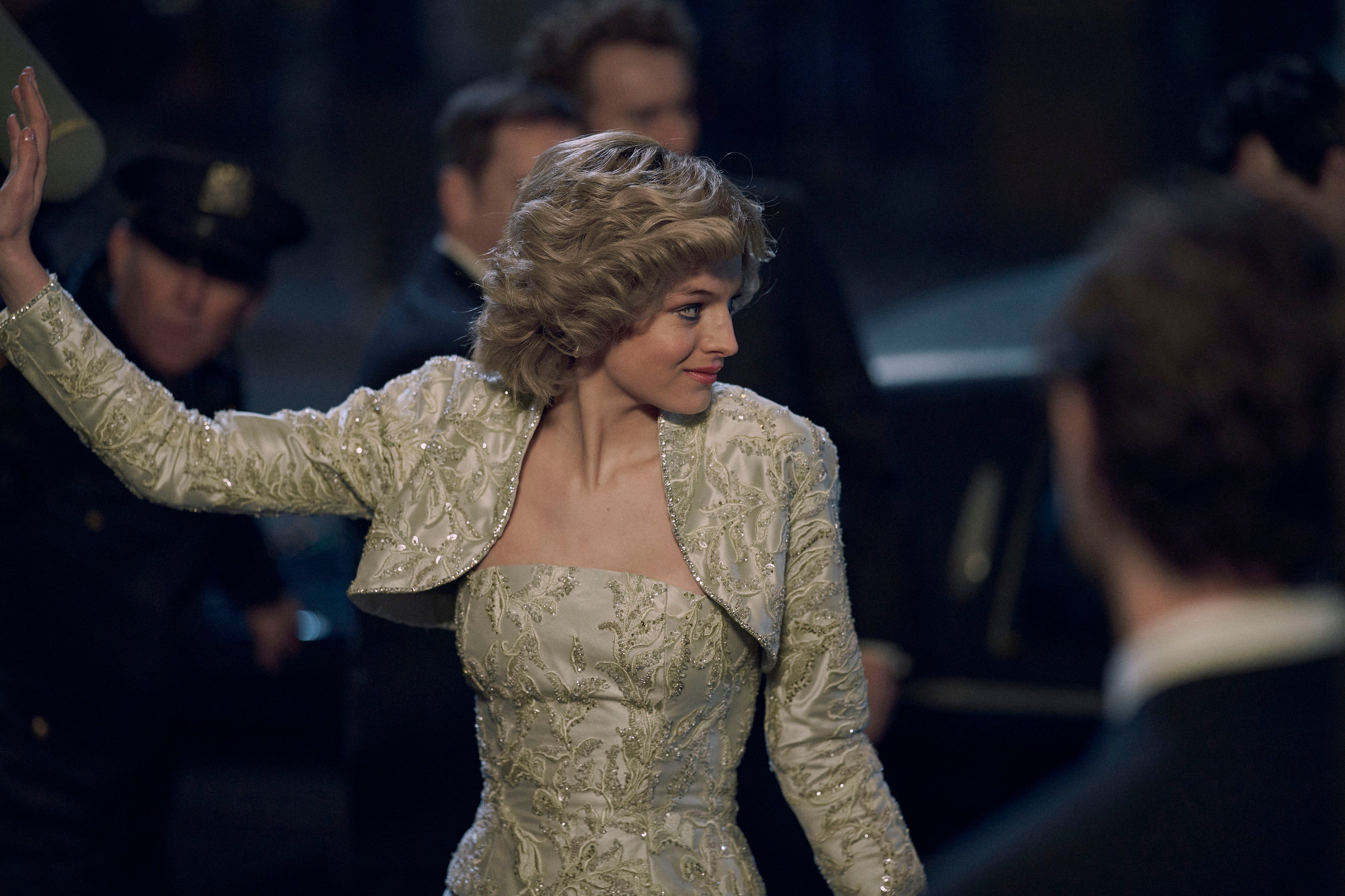 Emma Corrin as Diana in a gold brocade dress, waving with her right arm and looking to her left as she makes her entrance at an event, with security in the background