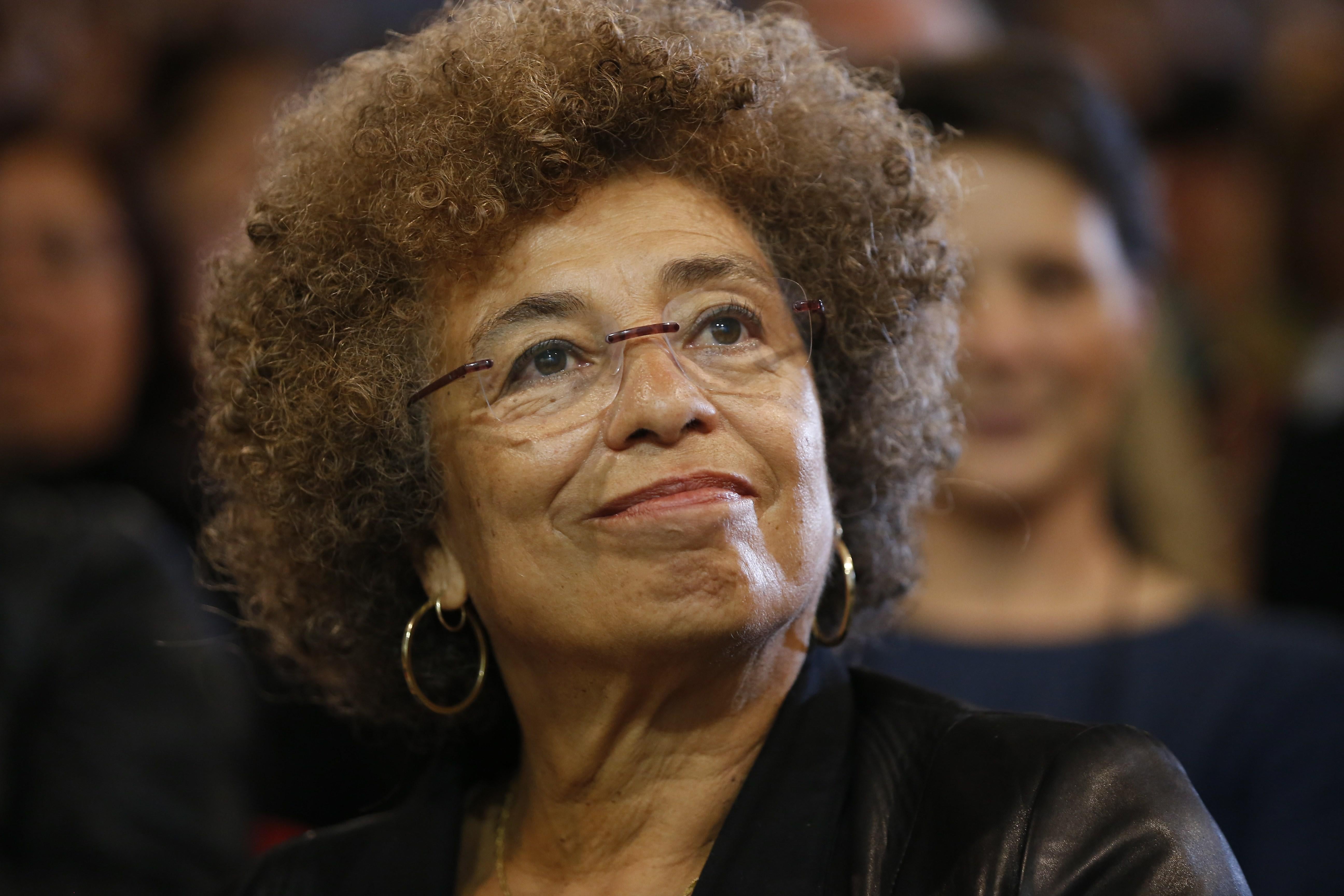 Angela Davis, sitting in front of a crowd, smiles toward the camera