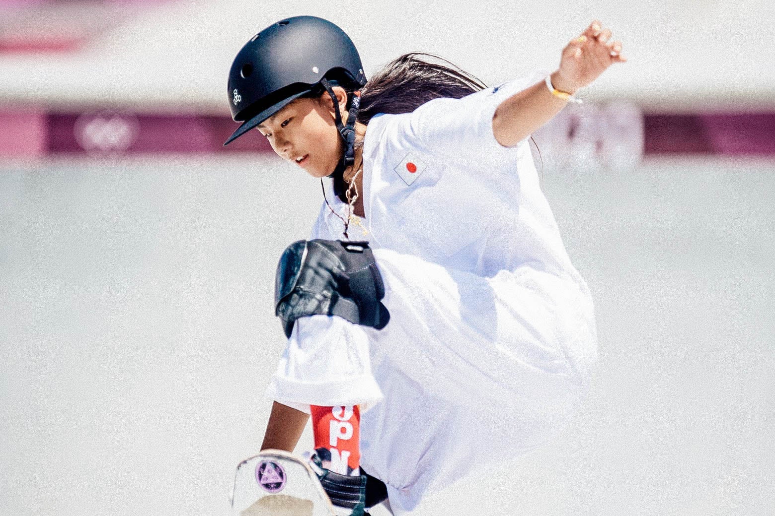 Hiraki on her skateboard ascending the park ramp, looking at her feet, left arm outstretched, in knee pads and helmet