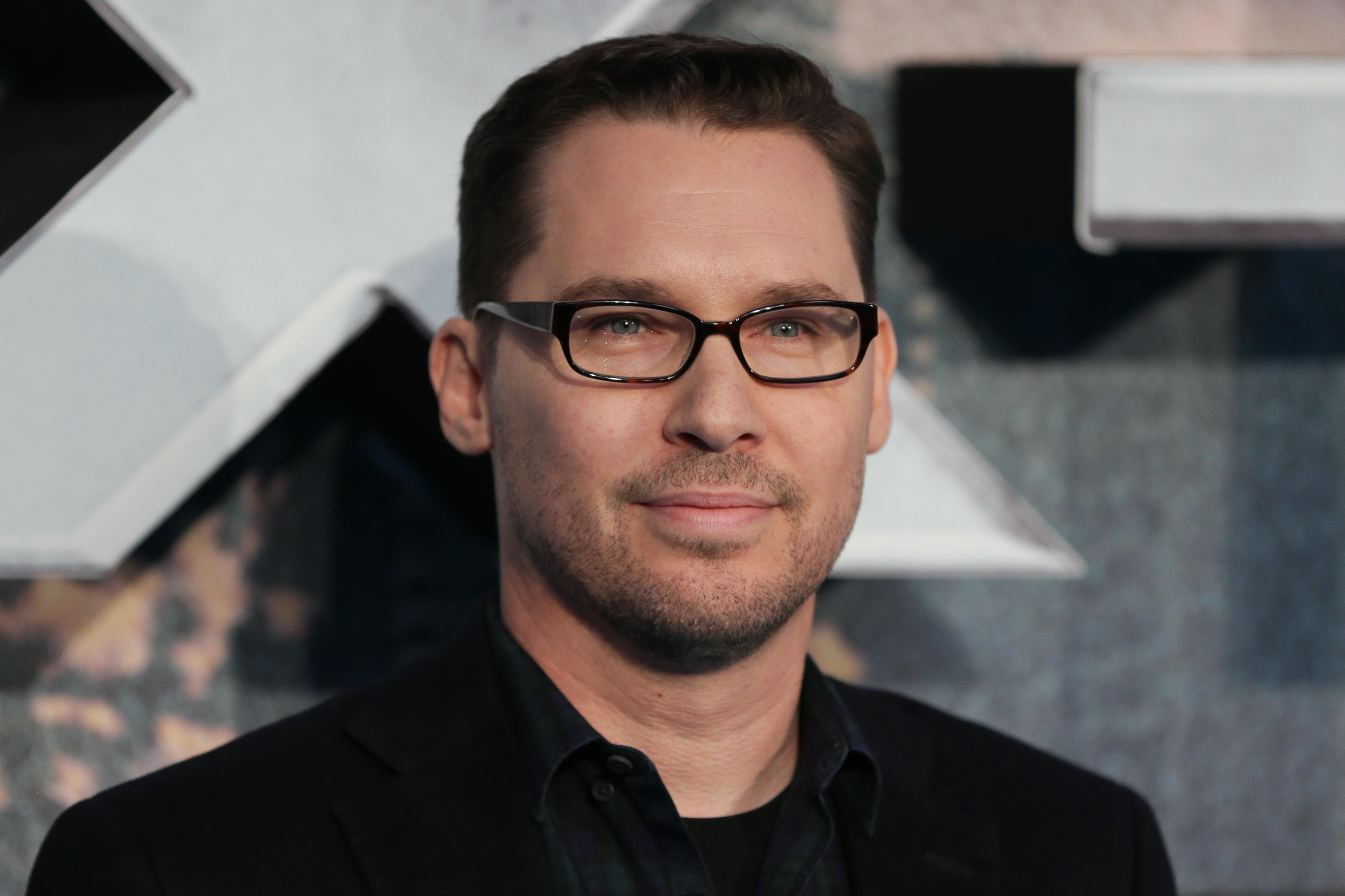 Bryan Singer, wearing a black suit and black glasses, stands in front of a large silver X.