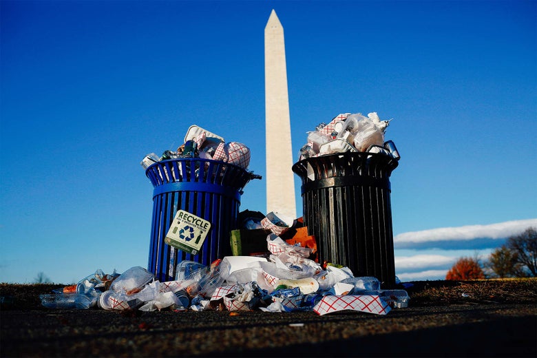 Trash overflows from a trash bin and a recycle bin, in front of the Washington Monument.