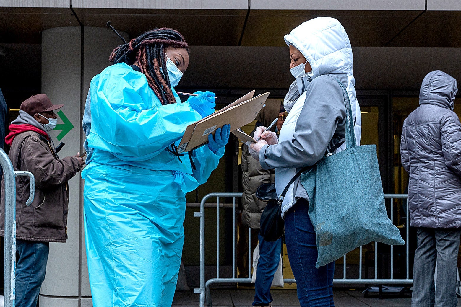 A woman in protect gear writes something on a clipboard next to a woman in a surgical mask.