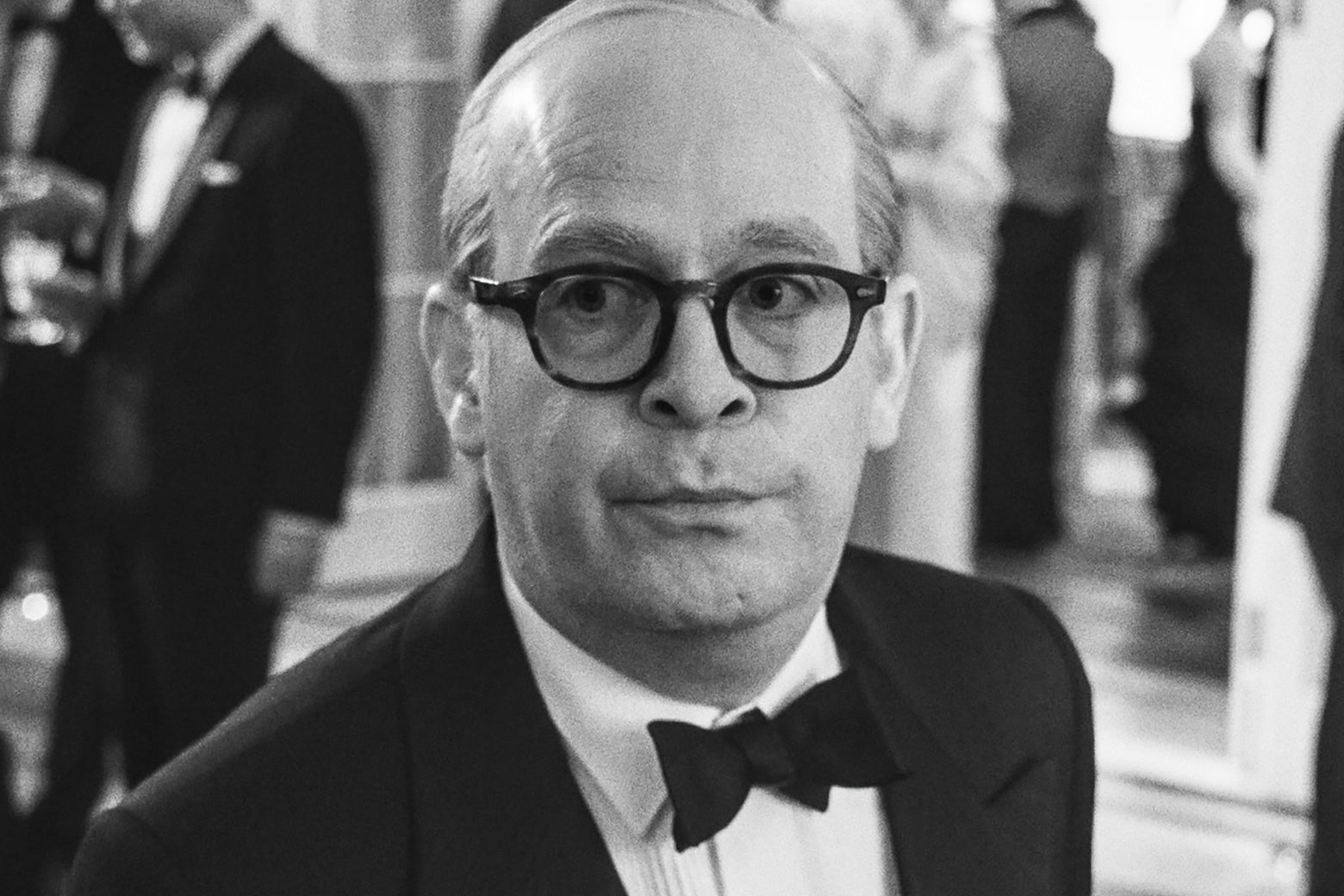 Tom Hollander as Truman Capote, wearing glasses and a tuxedo, in black and white.
