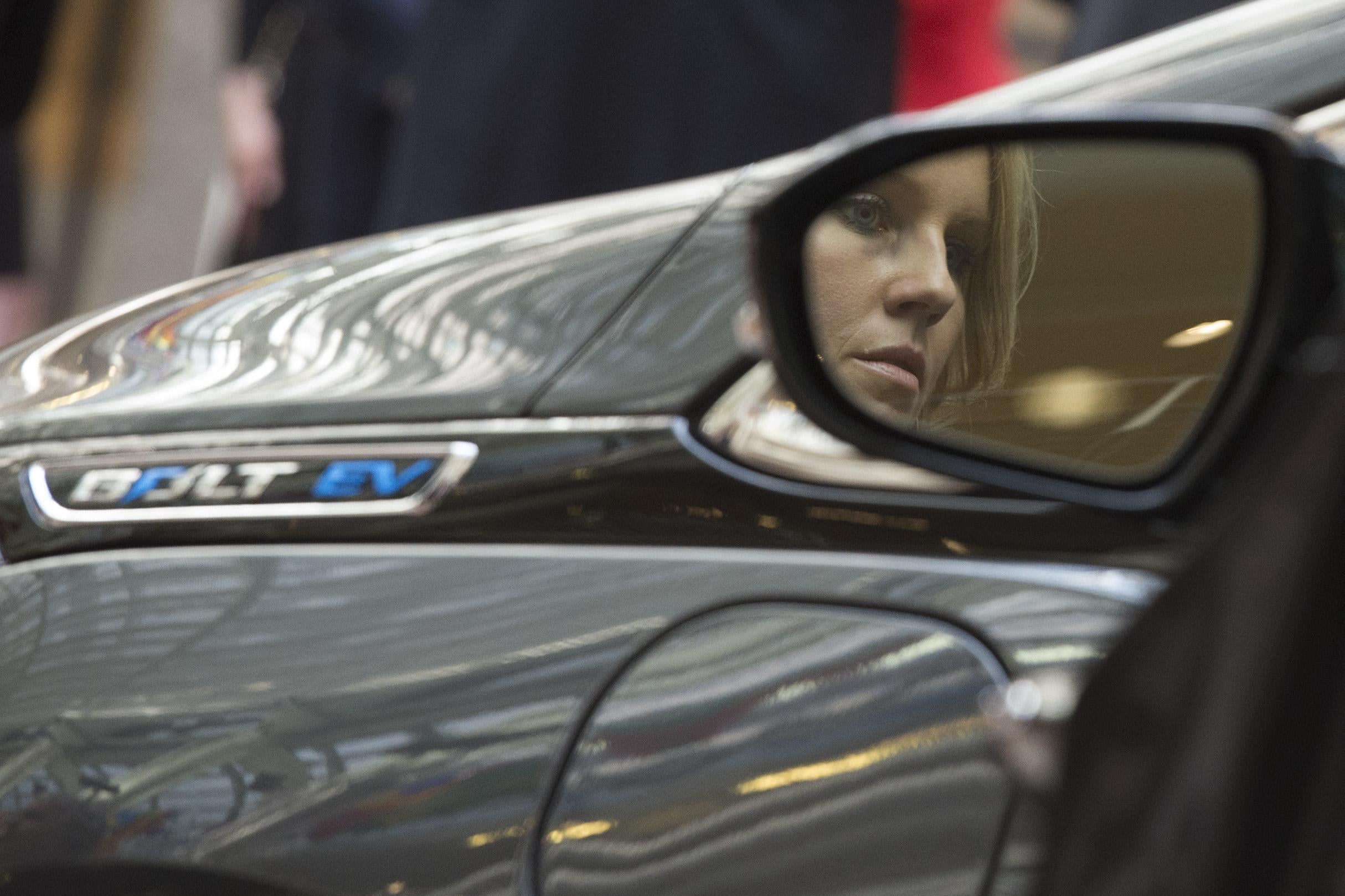 A woman's face appears in a driver's side mirror, against a Chevy Bolt in the background.t.
