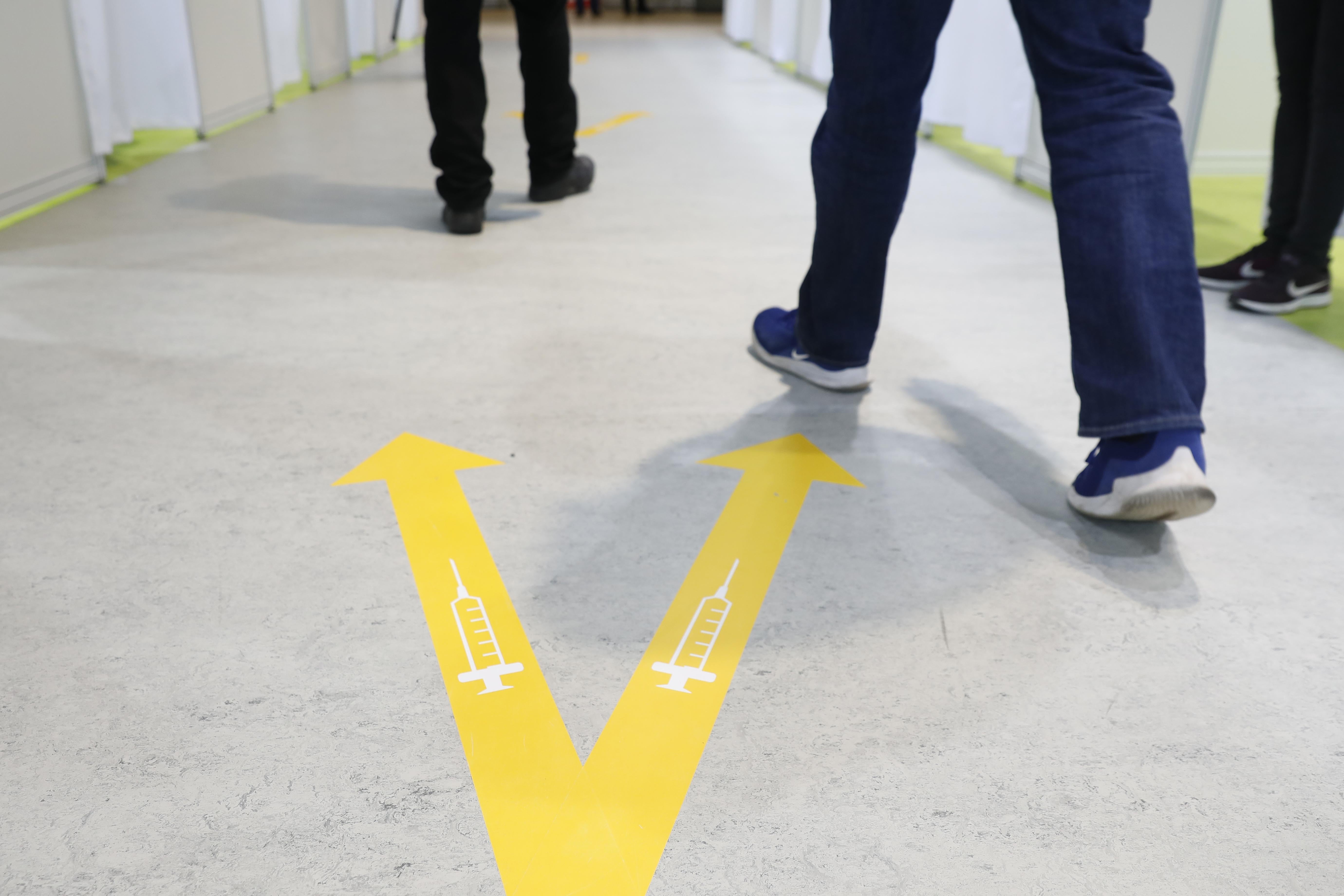 People's feet beside a yellow V painted on the floor, with a syringe in each arm of the V.