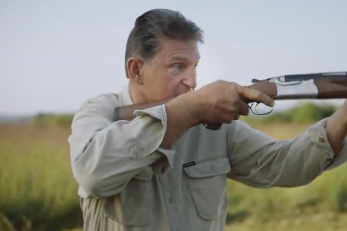 Manchin, standing in a field, levels and aims a shotgun.