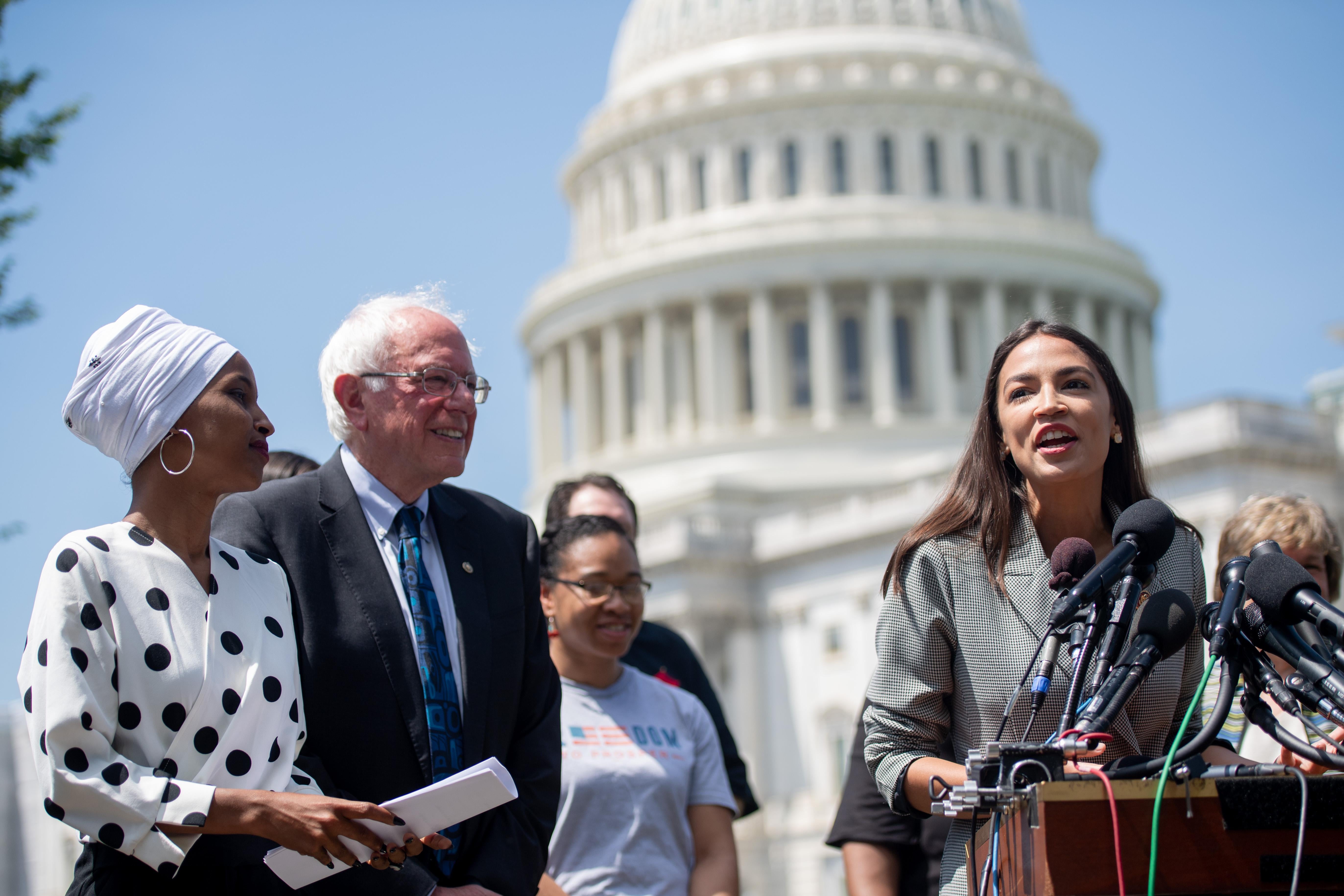 Ilhan Omar and Bernie Sanders look on as Alexandria Ocasio-Cortez speaks into several microphones at a podium. The Capitol dome and several other people are behind them.