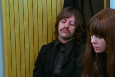 A woman with long brown hair stares darkly beside Ringo who's leaning his head against a paneled wall apparently asleep