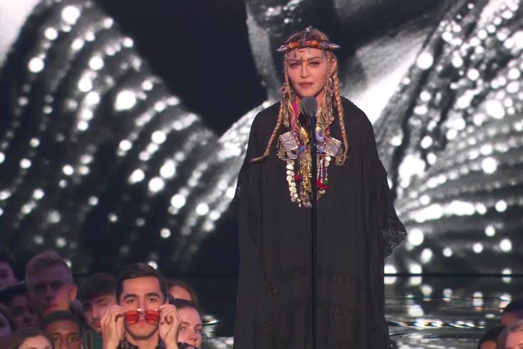 Madonna speaks into a microphone on stage at the VMA music awards, while a spectator puts on sunglasses.