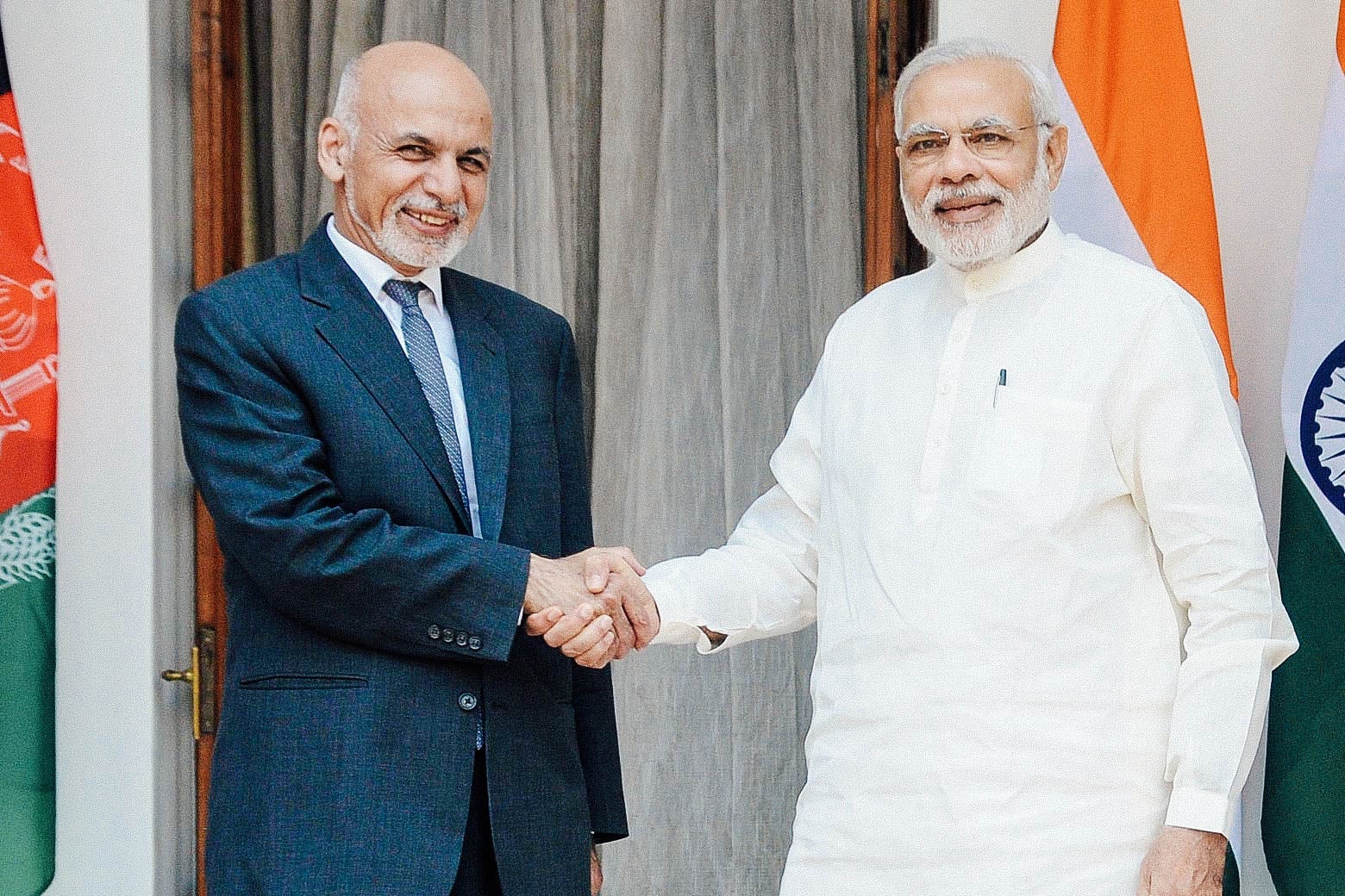 Two men shake hands and pose for the camera in front of flags for Afghanistan and India.
