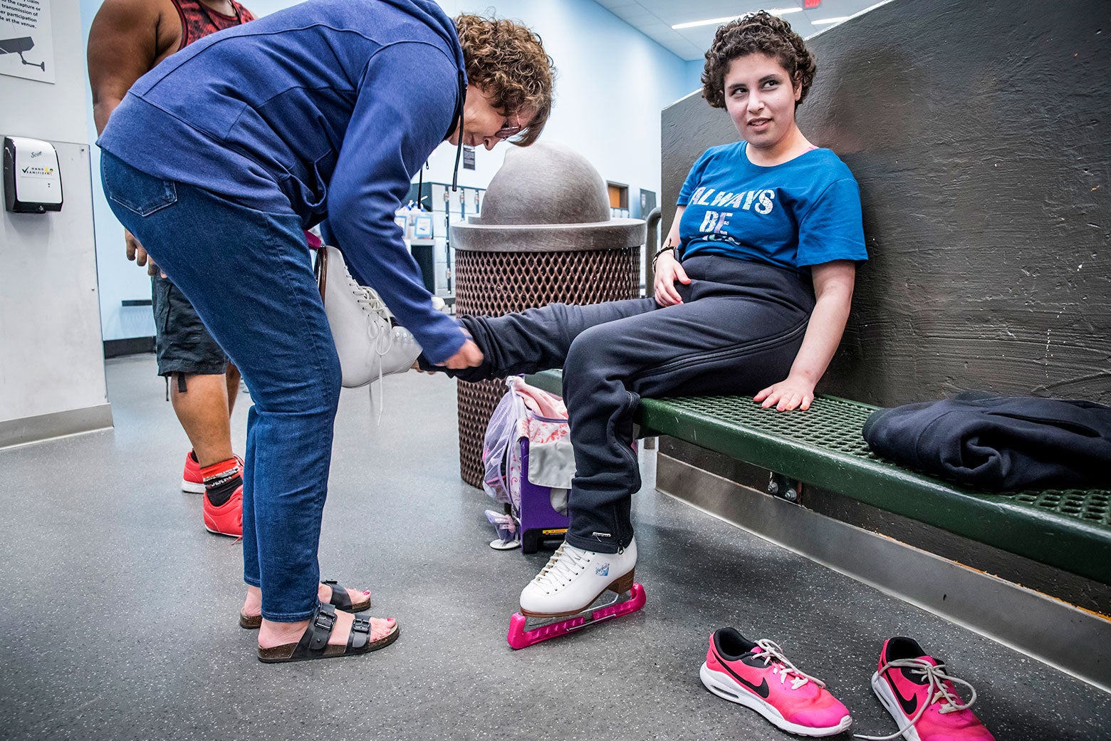 A woman sits on a bench, one foot in a skate on the ground as another woman standing in front of her ties the laces of her other skate.