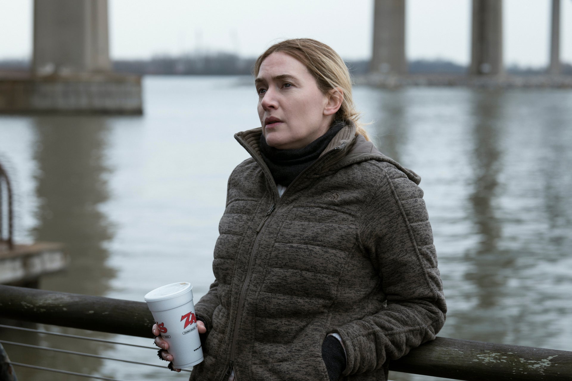 Kate Winslet as Mare looking pensive as she leans against a railing by the river. She is wearing a heavy coat and holding a takeout soda cup.