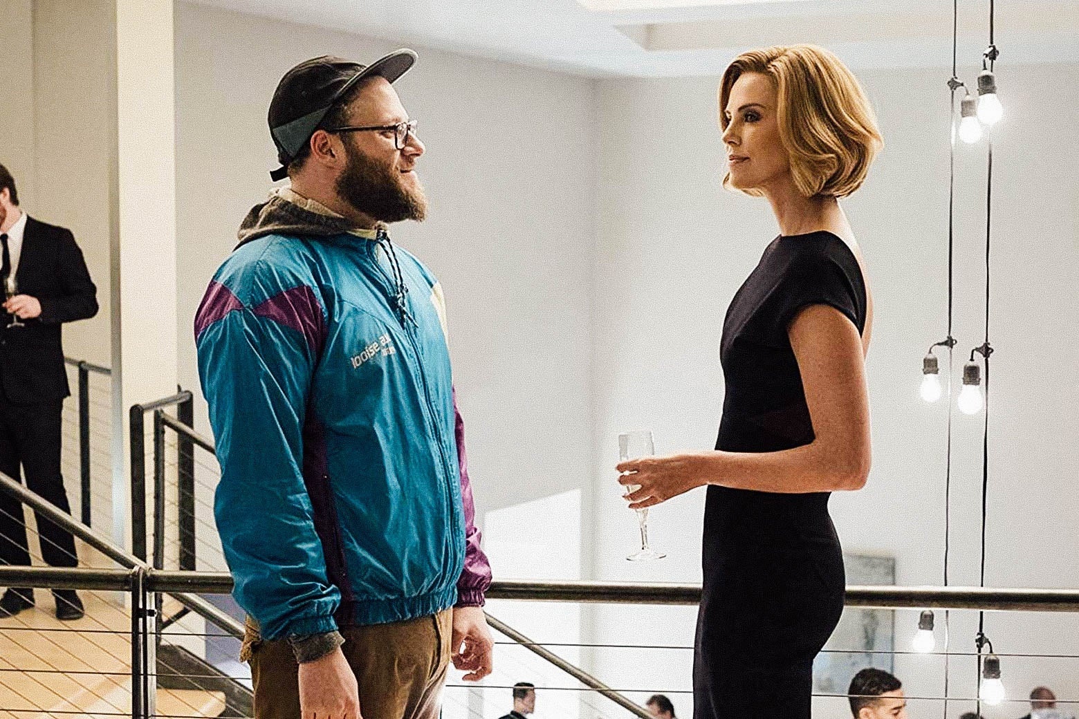 Seth Rogen wears a windbreaker and Charlize Theron wears a chic black dress as they talk at a party in a still from the movie.